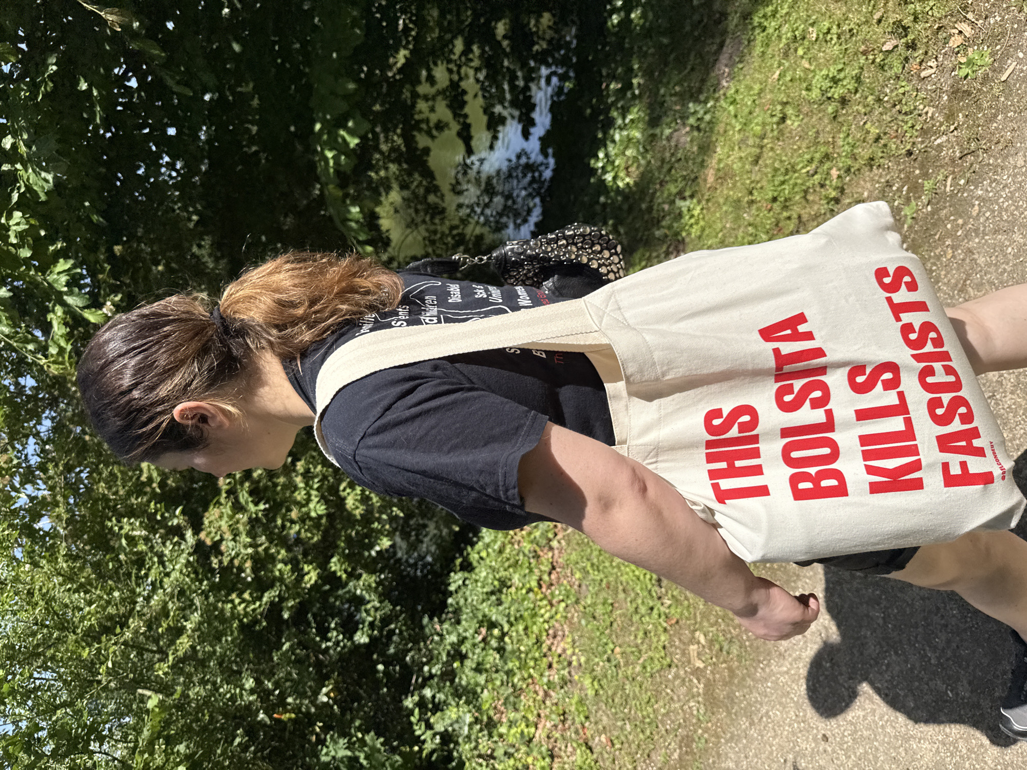 A person is walking on a path with trees and foliage visible in the background. The tote bag they are carrying is white and has red text that reads "THIS BOLSITA KILLS FASCISTS". They are wearing a black T-shirt that also has white text on it. The location appears to be a park or wooded area, and there is no specific indication that this photo was taken in Paris, France.