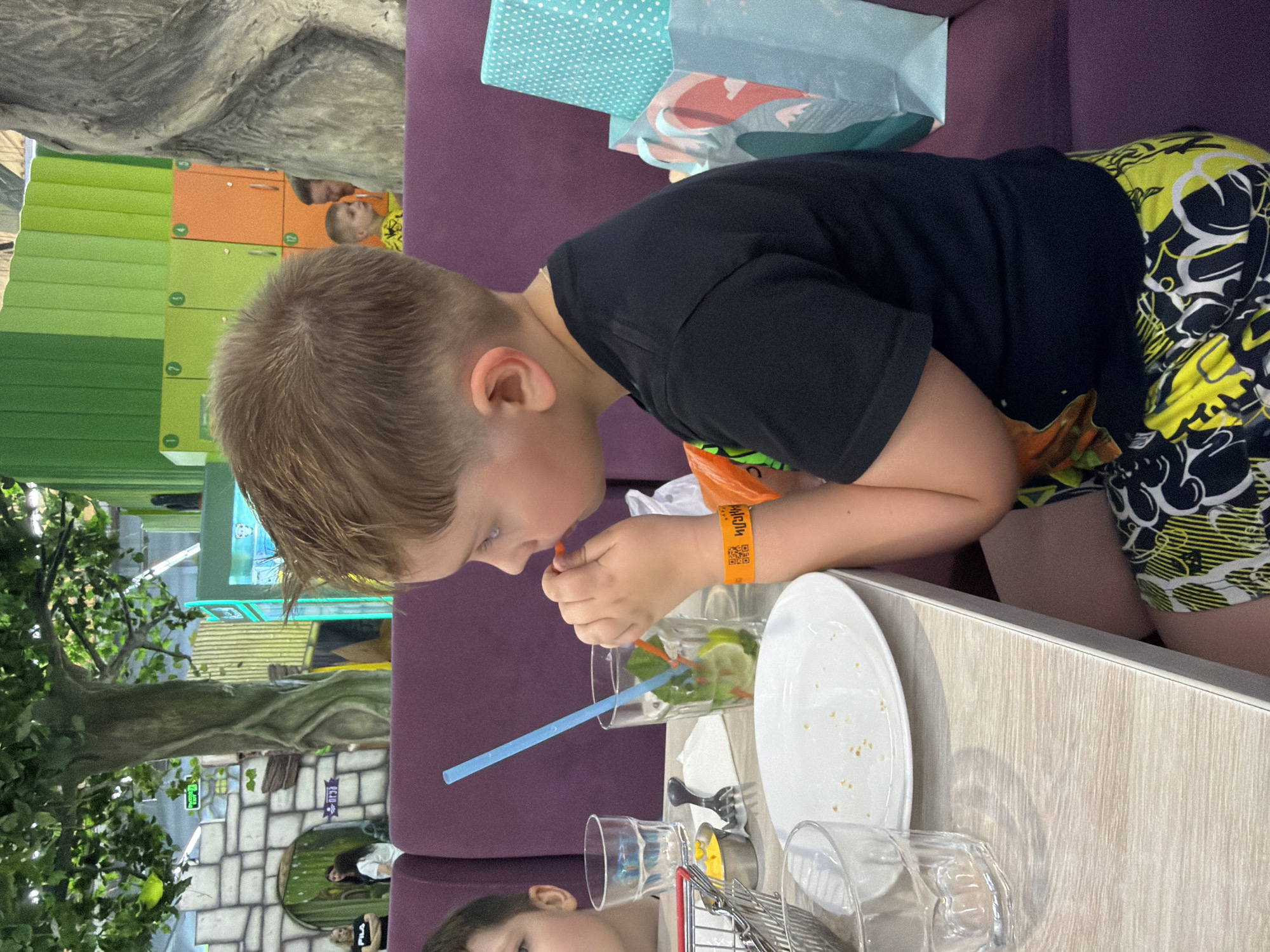 A young boy with blond hair is sitting at a table in a restaurant. He is wearing a black t-shirt and colorful shorts. He is drinking from a glass with a blue straw and a orange swizzle stick. There is a plate with crumbs on the table in front of him, and a glass of water to his left. In the background, there are several people, including a woman with dark hair wearing a white shirt, and another woman with dark hair wearing a black shirt. The restaurant appears to be decorated with a jungle theme, with artificial trees and plants. There are also colorful lockers in the background. The boy is wearing an orange wristband with a QR code on it.