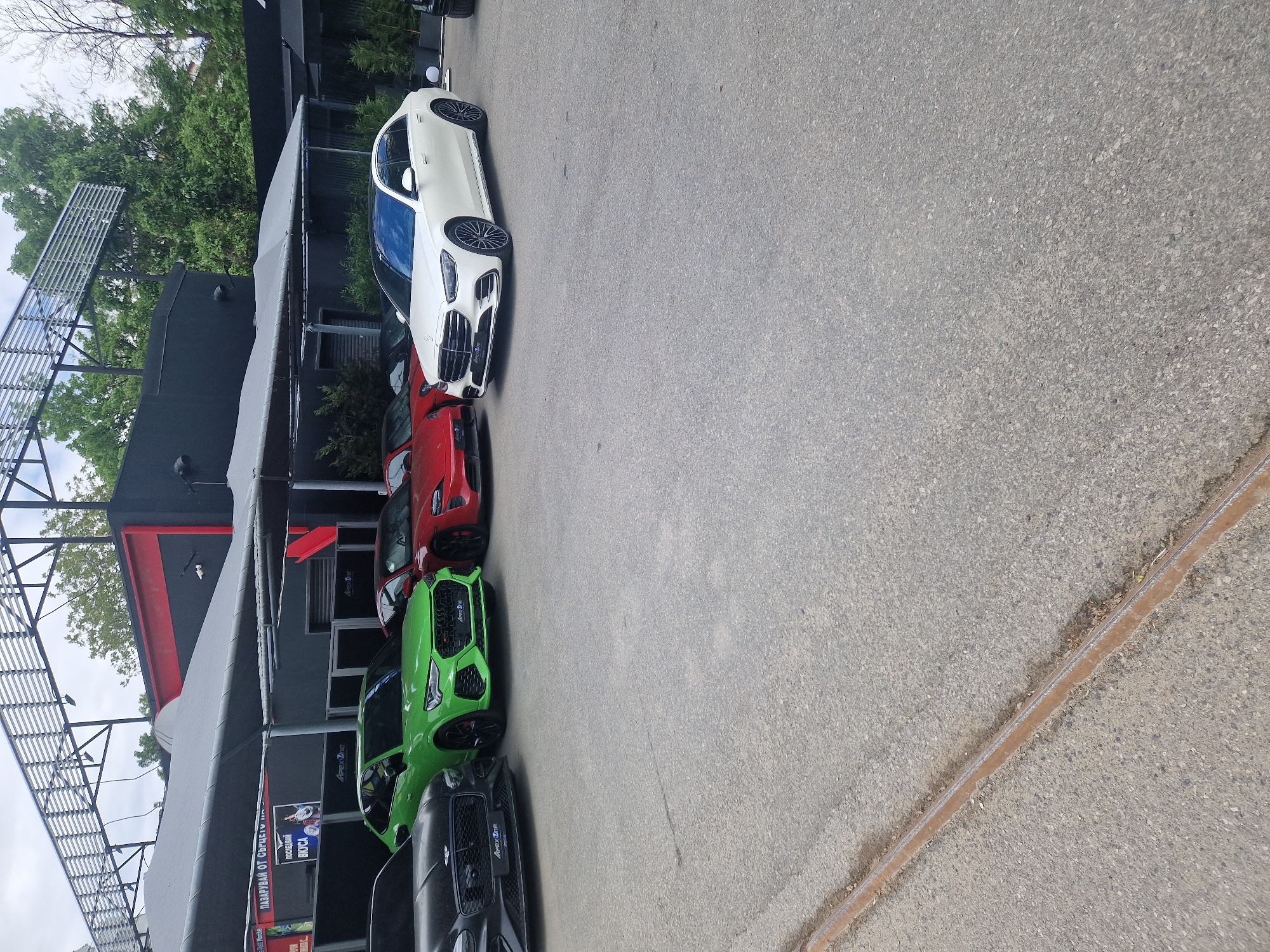 Here is a detailed description of the image:
**Scene:**
The image depicts a commercial area in what appears to be an urban environment. The scene is dominated by a line of parked luxury cars, which include a white sedan, a red sports car, a green sports car, and a black luxury car. These cars are parked adjacent to a building that features a modern, industrial design with a black facade and a gray awning. A metal staircase structure is visible on the left side of the building. Signage, including text in Bulgarian and English (e.g., "Apex Dine," "ПАЗАРУВАЙ ОТ СЪРЦЕТЕ," which translates to "Shop from the heart," "ПОСЛЕДВАЙ ВКУСА" which translates to "Follow the taste"), suggests that this is a commercial location, possibly a car dealership or showroom combined with a restaurant. The foreground is dominated by a gray asphalt pavement.
**Environment:**
The location is characterized by a mix of architectural elements and natural elements. Trees are visible in the background, adding a touch of greenery. The sky is visible above, with what appears to be a cloudy or overcast condition. The overall scene conveys a sense of urban commerce and luxury.