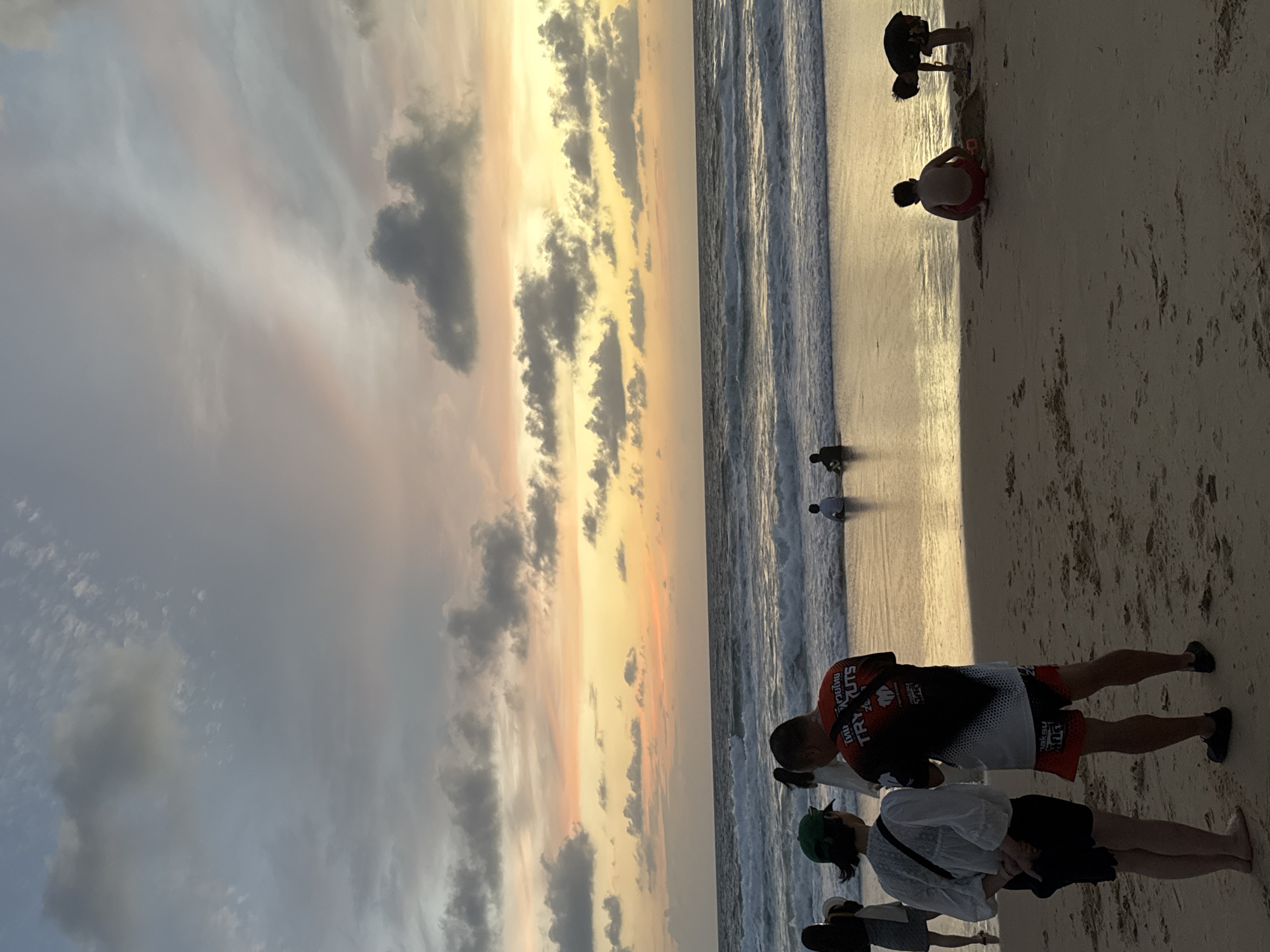 At sunset on Karon Beach in Karon, Thailand, several people are present. In the foreground, a woman wearing a green hat and a white shirt stands facing a man in a red and black t-shirt and red shorts. To the right, two young boys are playing in the sand. One boy is squatting, and the other is digging with a shovel. Further out in the ocean, a few individuals are visible in the water. The sky displays a mix of grey clouds and warm, pastel hues of orange and yellow, indicating the sun is setting. The waves are gently rolling onto the sandy beach.
