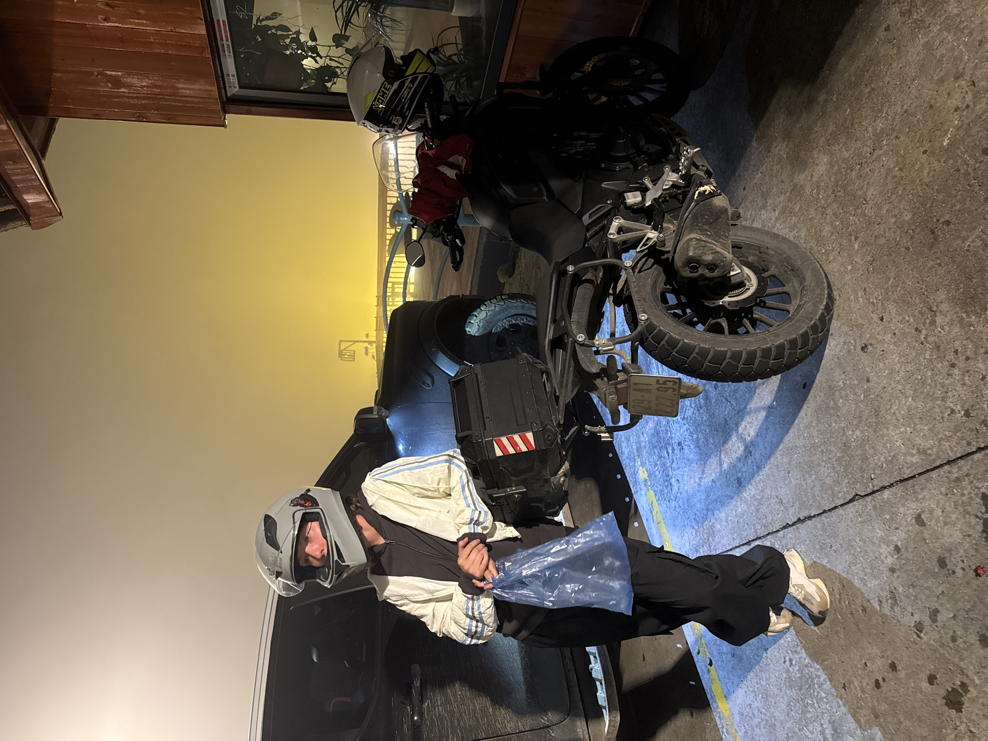 A person wearing a motorcycle helmet and a blue and white jacket stands next to a parked black motorcycle. The motorcycle is facing to the right and has a white and yellow helmet resting on its seat. The scene appears to be in an urban area, possibly Binh Lu, Vietnam, based on the license plate of the motorcycle. The setting has yellow lighting which could indicate night or dusk. The person stands near a car. The ground is concrete.
