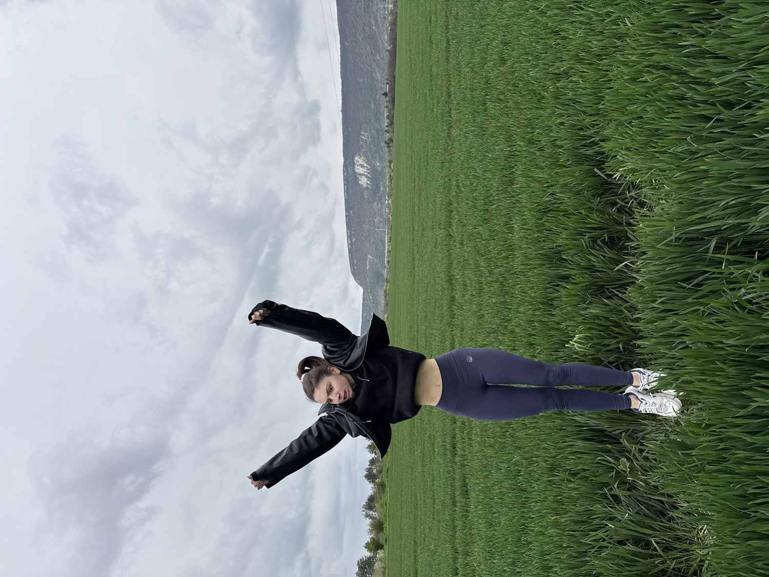 An upright image shows a young woman posing in a vast field of vibrant green grass under an overcast sky in Shumen, Bulgaria. The woman is centered in the frame, extending her arms outwards and slightly upwards. She has dark hair tied in a ponytail and is looking towards the viewer with a calm or slightly smiling expression.
She is dressed in a black long-sleeved jacket, dark blue form-fitting leggings, and white athletic shoes. A small, white, circular logo, resembling a stylized letter (possibly an 'A'), is visible on the left hip of her leggings. The field of green plants, likely a crop like wheat or barley, stretches into the distance. In the background, rolling hills or mountains are visible, with some sparse tree lines along their slopes. The sky is filled with grey clouds, indicating an overcast day. The setting suggests a rural or natural outdoor environment during daytime.