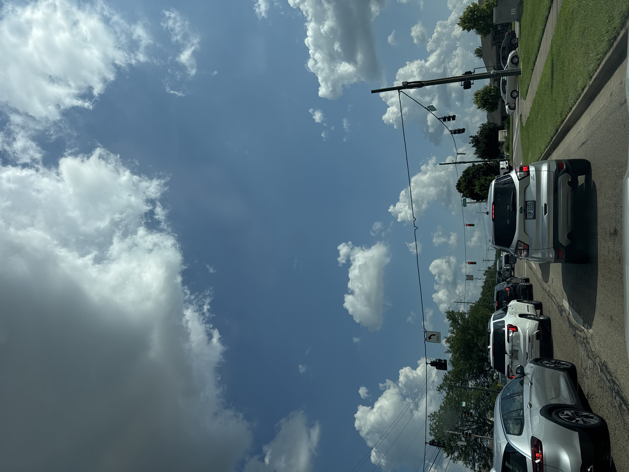 The image shows a traffic scene in Dayton, United States, likely on a bright, partly cloudy day. Several vehicles are stopped on a paved road, suggesting a traffic light or congestion. The vehicles include a silver SUV in the foreground, a white SUV behind it, and other cars further down the road. Traffic lights are visible, some showing red. Trees line the side of the road, and there are buildings and grassy areas in the background. The sky is a mix of blue with large, fluffy white and gray clouds. There are power lines and poles visible. No people are clearly visible. The scene appears to be an intersection or a stretch of road with multiple lanes.