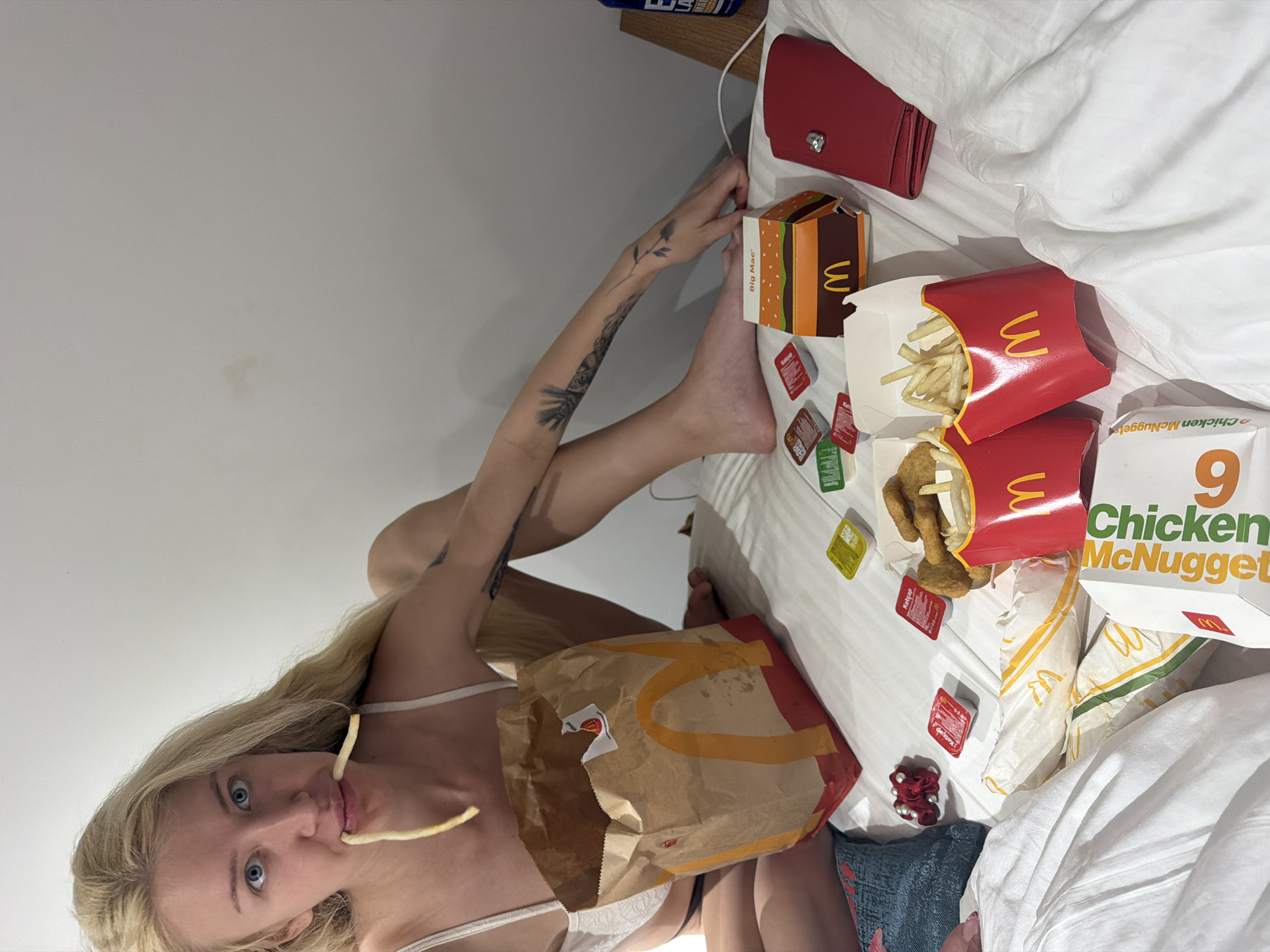 A Caucasian female with long blonde hair and blue eyes is lying on a white bed in a room. She is eating a french fry. A brown McDonald's bag is on her body. Scattered around her on the bed are McDonald's food items, including french fry containers, a box of chicken McNuggets, a Big Mac box, and various condiment packets. A red purse is also on the bed. Part of a wooden nightstand and blue book can be seen in the background. The location is Küçükçekmece, Turkey.