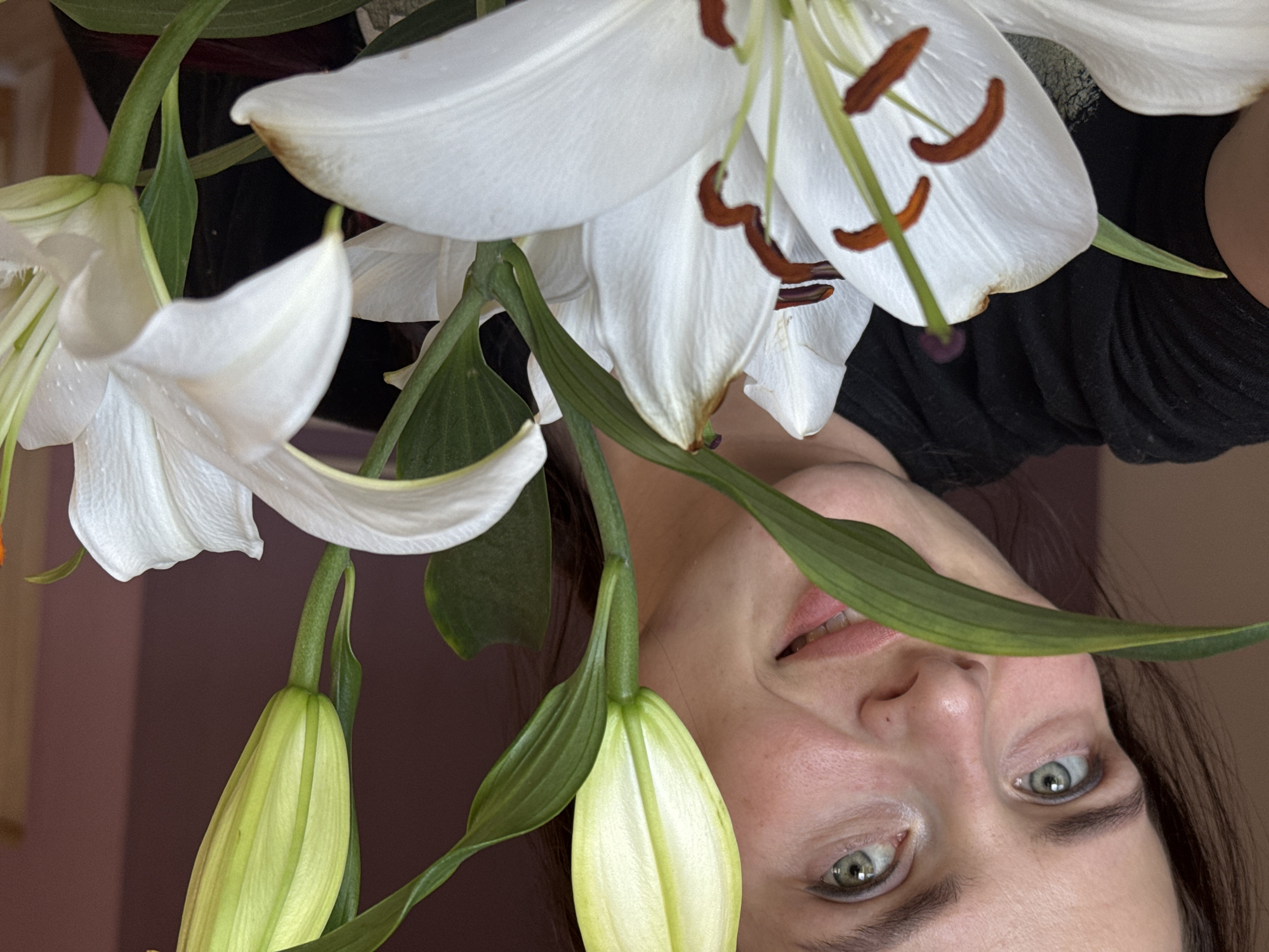 A close-up, upside-down shot features a woman and white lilies in what appears to be an indoor setting in Rusalevka, Russia. The woman's face is partially visible, with her green eyes looking towards the camera and her mouth slightly open in a smile. She has dark hair and is wearing a black top. The lilies are in various stages of bloom, with some fully open and others still in bud. Their white petals and green stems dominate the foreground. The background is a muted brown color, suggesting a wall or fabric. The lighting is soft, indicating it might be daytime, but no specific time can be determined. There are no visible text elements in the image.