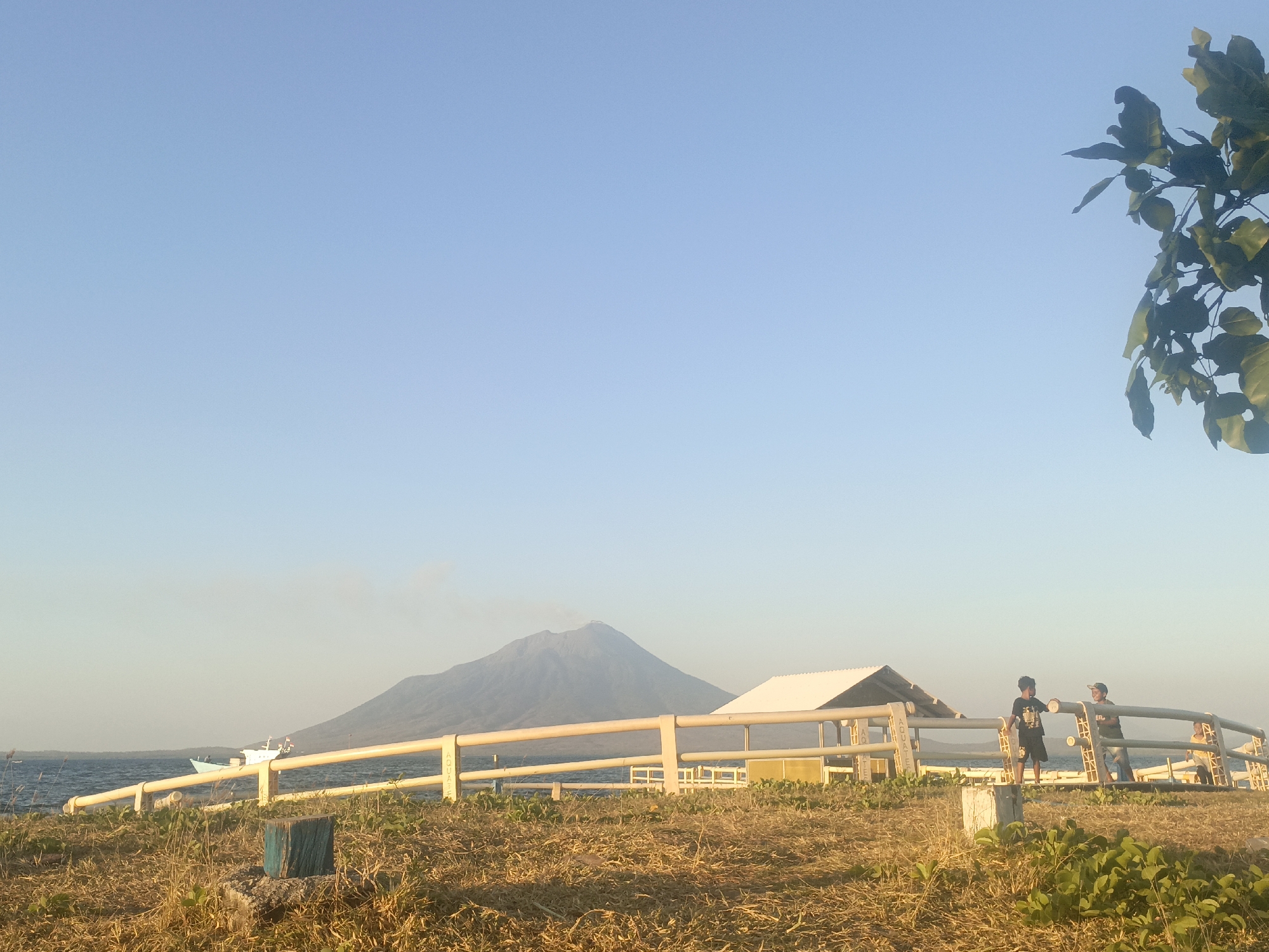 In the city of Lewoleba, Indonesia, this outdoor scene captures a coastal view with a majestic volcano in the distance. The foreground is a dry, grassy field with some green vegetation at the bottom. A yellow, slatted fence runs horizontally across the mid-ground. Two young males are standing near the fence, one in a black t-shirt and shorts, the other in a green t-shirt and dark shorts, appearing to interact. Behind the fence, a simple open-sided structure with a white roof is visible. In the background, the calm, dark blue sea stretches out, with a light blue boat and a white boat with red accents on the water. The imposing volcano, with a plume of smoke rising from its summit, dominates the far background under a clear, pale blue sky. The sun appears to be low in the sky, casting a warm light on the scene.