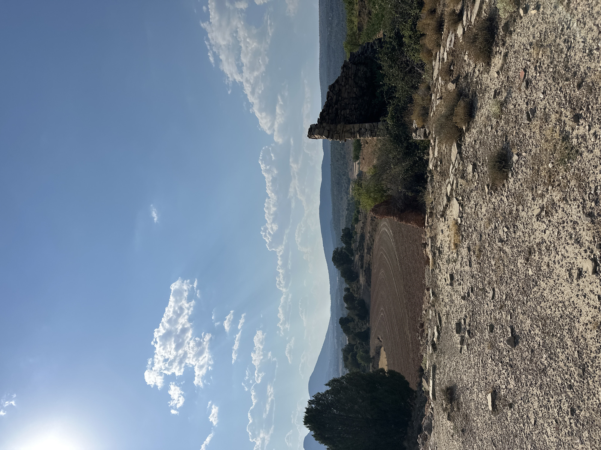 The image depicts a sunlit, rural landscape under a bright blue sky dotted with white clouds. In the foreground, the ground is covered with gravel, stones, and scattered flat slabs, suggesting remnants of a paved surface or foundation. A partially ruined stone structure stands prominently on the right side of the frame, its weathered stones forming a jagged silhouette against the sky. To the left, a large, dark green evergreen tree dominates the mid-ground. Beyond the tree, a brown, plowed field is visible with curved furrows, indicating agricultural activity. The background is characterized by rolling hills and mountains, with vegetation appearing sparse in some areas and more dense in others. The overall scene suggests an arid or semi-arid environment. The location is identified as Alpuente, Spain. No people or animals are visible in the image.
