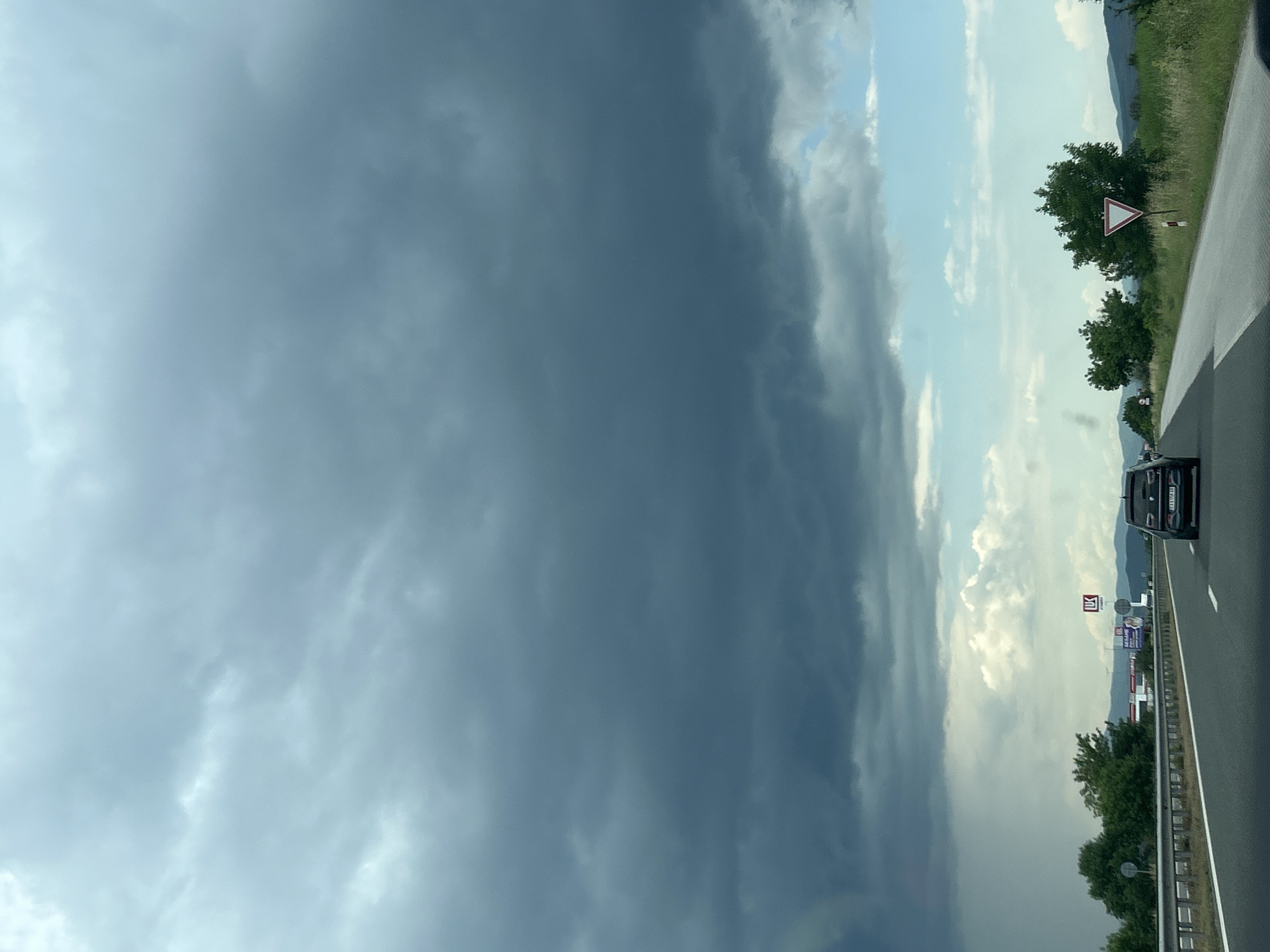 The image depicts a highway scene in Shtarkovo, Bulgaria, under a sky largely covered by dark, heavy stratocumulus clouds, indicative of an impending or ongoing storm. The upper two-thirds of the frame are dominated by these grey clouds, with a discernible curtain of rain falling on the far left. Towards the lower right, the sky transitions to a lighter blue with scattered white clouds. The perspective is from within a vehicle traveling on a multi-lane asphalt highway, marked with white lines. A metal guardrail borders the left side of the road, beyond which are green trees and distant, hazy mountains. On the right side of the highway, a grassy verge leads to a "Give Way" (yield) sign (red border, white inverted triangle) and a cluster of green trees. In the mid-ground on the right, advertising signs, including one with an "IK" logo and another with "SASAME" text, are visible near structures that appear to be roadside businesses. A black SUV, bearing a partially visible Bulgarian license plate (e.g., "CA 6160 AP"), travels ahead in the same lane. No individuals are visible. The environment suggests an open rural or semi-urban area, with mountains in the far distance on both sides.