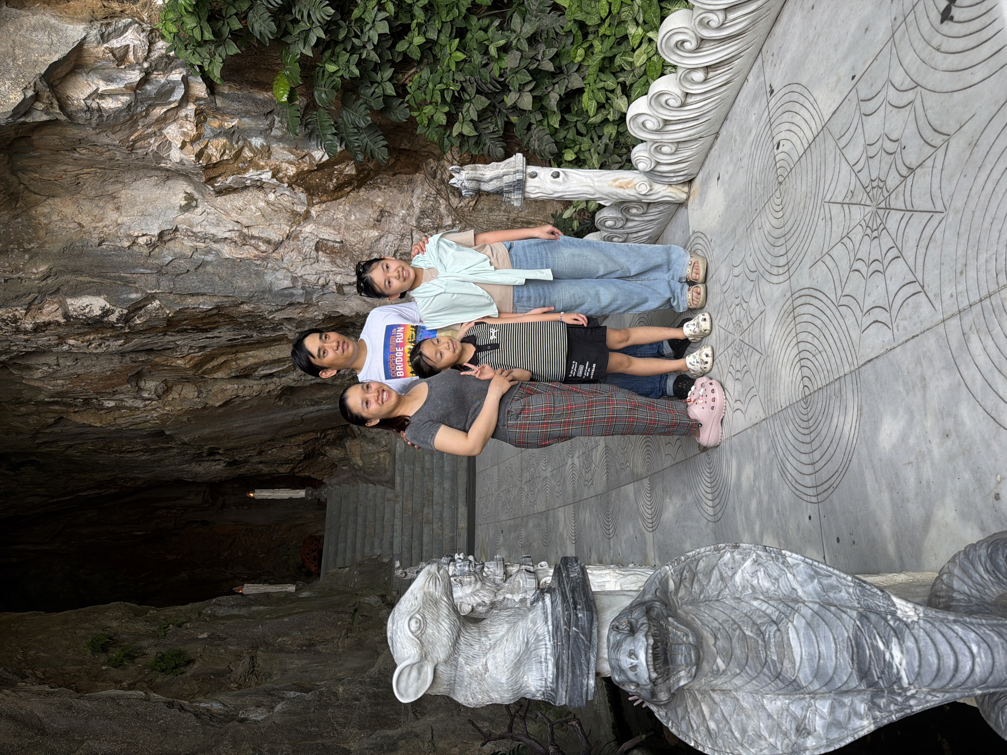 Four people are arranged in a row for a photograph. The lowest person is a woman wearing a grey shirt and red plaid pants. Above her is a male wearing a white shirt with a book on the front. Next is a girl in a striped shirt and black skirt, and above her is a girl wearing blue jeans and a light blue shirt. They are positioned next to decorative gray marble statues of a rabbit and a snake. They are in front of the entrance to a cave with steps leading inside. The cave entrance is in a rock face with green foliage above. The pavement in front has etched spider web designs. This photograph was taken at the Marble Mountains in Đà Nẵng, Vietnam.
