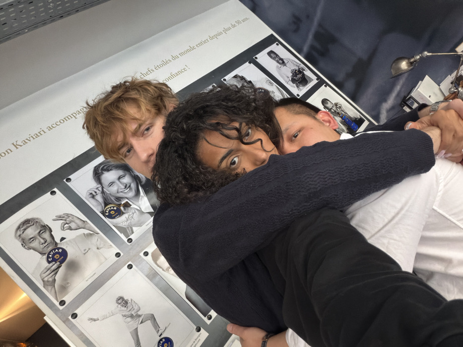 Three individuals are captured in a close-up, diagonally oriented portrait, appearing to be in a tight embrace or posing for a group photograph. The topmost person is a male with light brown hair and fair skin, looking directly into the camera. Below him, a female with dark curly hair and dark skin occupies the central frame, wearing a dark textured long-sleeved top and dark pants. Her right arm is wrapped around the third individual, and she is also looking directly at the camera. Partially visible beneath the female is a male with short light brown hair, showing the side of his face, a white shirt, and his left hand, adorned with a watch, grasping the female's right hand.
The background consists of a wall displaying multiple framed black and white portraits of chefs, each holding a blue and gold tin marked "CAVIAR." Visible French text on the wall includes phrases such as "KAVIARI accompag..." and "chefs étoilés du monde entier depuis plus de 50 ans." A metallic, articulated desk lamp is partially visible in the lower right corner of the frame. Based on the prominent "KAVIARI" branding and French text, the scene is located at the Kaviari boutique or headquarters, specifically at 13 Rue de l'Arsenal, 75004 Paris, France.