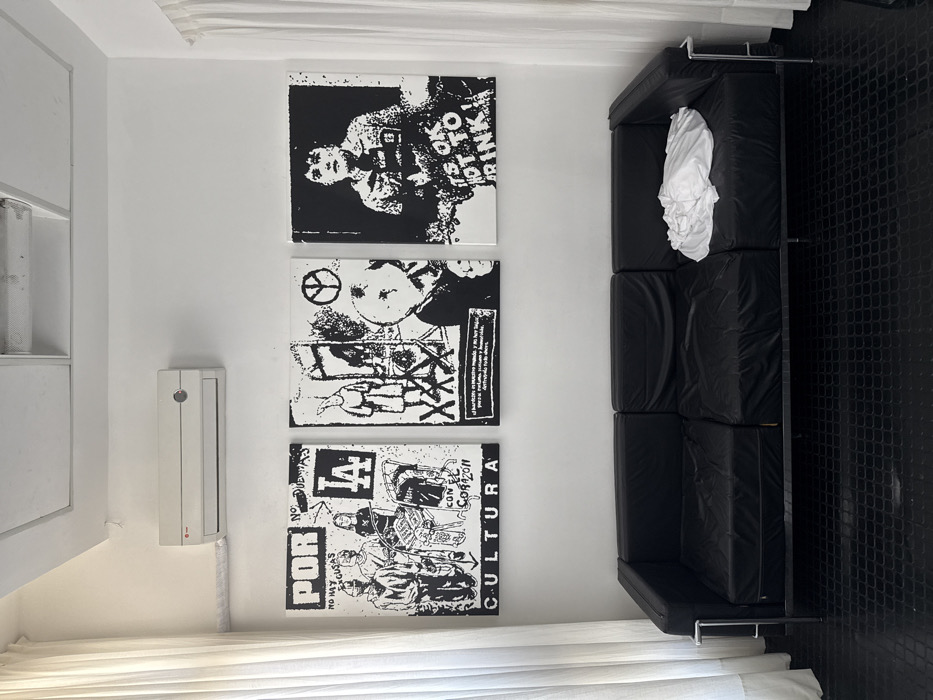 A white room has three black-and-white art pieces stacked vertically on the wall, each appearing to be collage or print artwork. Beside the artwork is a long, modern black couch with chrome legs. On the couch rests a white blanket or sheet. An intercom system is mounted on the wall adjacent to a built-in white cabinet. A white curtain stretches along the bottom. The floor is a black, textured tile.