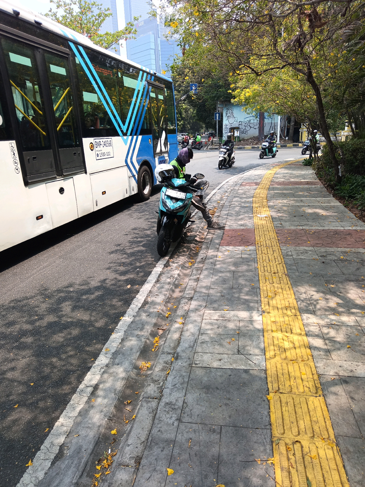 The image depicts a street scene in Central Jakarta, Indonesia, on a sunny day. A white and blue bus, identified by the license plate "BMP-240368", is parked on the left side of the road, partially obscuring the view of the sidewalk. The bus has blue geometric patterns on its side and yellow interior bars visible through the windows.
In the foreground, a turquoise scooter is parked on the sidewalk. A person wearing a helmet and a green jacket is seated on the scooter, with their legs extended. Yellow tactile paving lines the edge of the sidewalk, separating it from the grey paved area. Fallen yellow leaves are scattered across the sidewalk and the road.
Further down the street, several other scooters, ridden by people wearing helmets, are visible on the road. The background features trees with yellow flowers and tall buildings, one of which has "UOB" visible on it. The overall environment suggests a busy urban setting with moderate traffic.