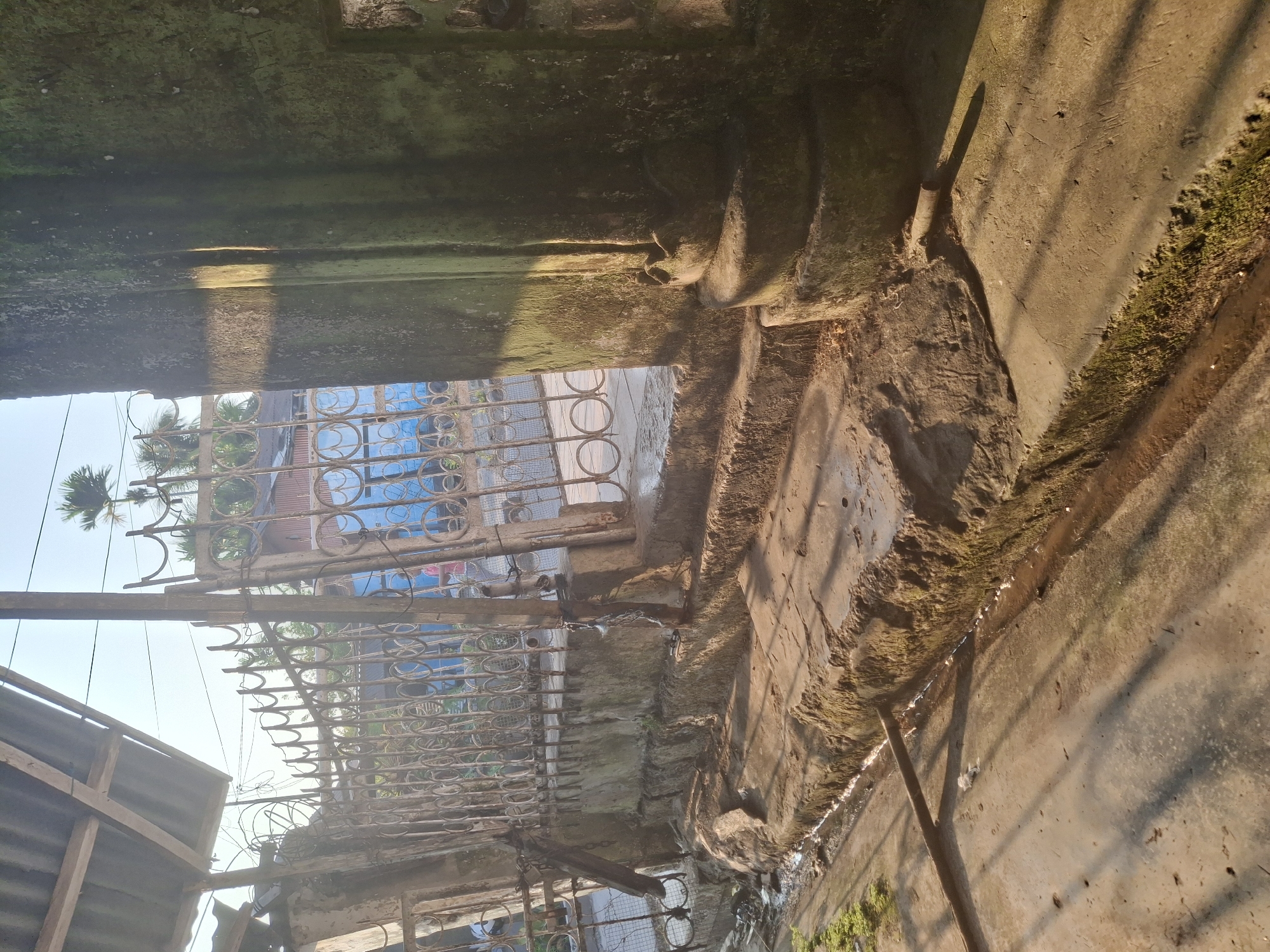 The image shows a sunlit exterior scene, possibly in Biak, Indonesia, featuring a weathered, rusty metal gate with circular patterns. To the right of the gate is a large, moss-covered stone pillar with decorative carvings at its base. The ground in front of the gate consists of rough-hewn stone steps leading down to a concrete pathway. A narrow channel of water runs along the edge of the steps. Behind the gate, a blue structure with vertical paneling and a metal roof is visible, along with palm trees in the distance. The overall impression is one of age and disrepair, with strong shadows cast by the sunlight.