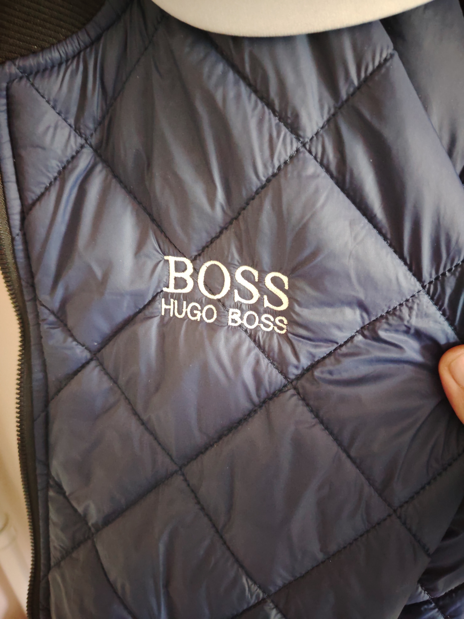 The image displays a close-up view of a navy blue quilted garment, likely a vest or jacket, featuring a diamond stitching pattern across its surface. Centered on the garment, two lines of white embroidered text are visible. The upper line reads "BOSS" in large, sans-serif capital letters. Directly beneath it, the second line reads "HUGO BOSS" in smaller, sans-serif capital letters. The fabric exhibits a slight sheen and some minor creasing. A black zipper is visible running vertically along the left edge of the garment. A portion of a white or light-colored curved object, possibly a garment hanger, is present at the top of the frame. Along the right edge of the image, a human thumb and the tip of an index finger are visible, resting on or gripping the garment. No other environmental or background details are discernible. The image provides no information regarding its specific location in Huntingdonshire, United Kingdom, or any other geographical area.