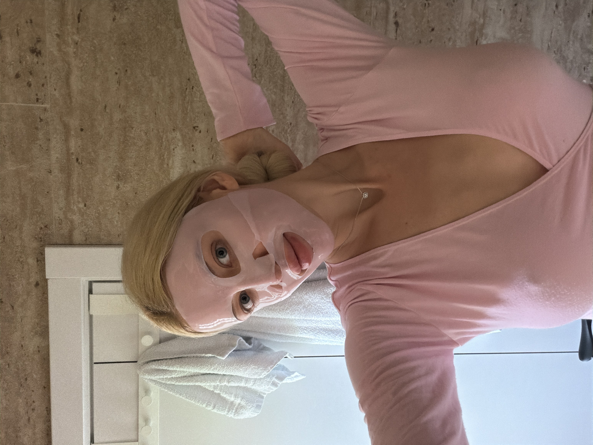 A woman with blond hair pulled back in a bun is wearing a pink long-sleeved shirt and a pink sheet face mask. She is sticking her tongue out slightly. She is wearing a necklace. A white cabinet with a towel is behind her. The wall behind the cabinet is beige and appears to be made of tile. The location is not determinable from the image.