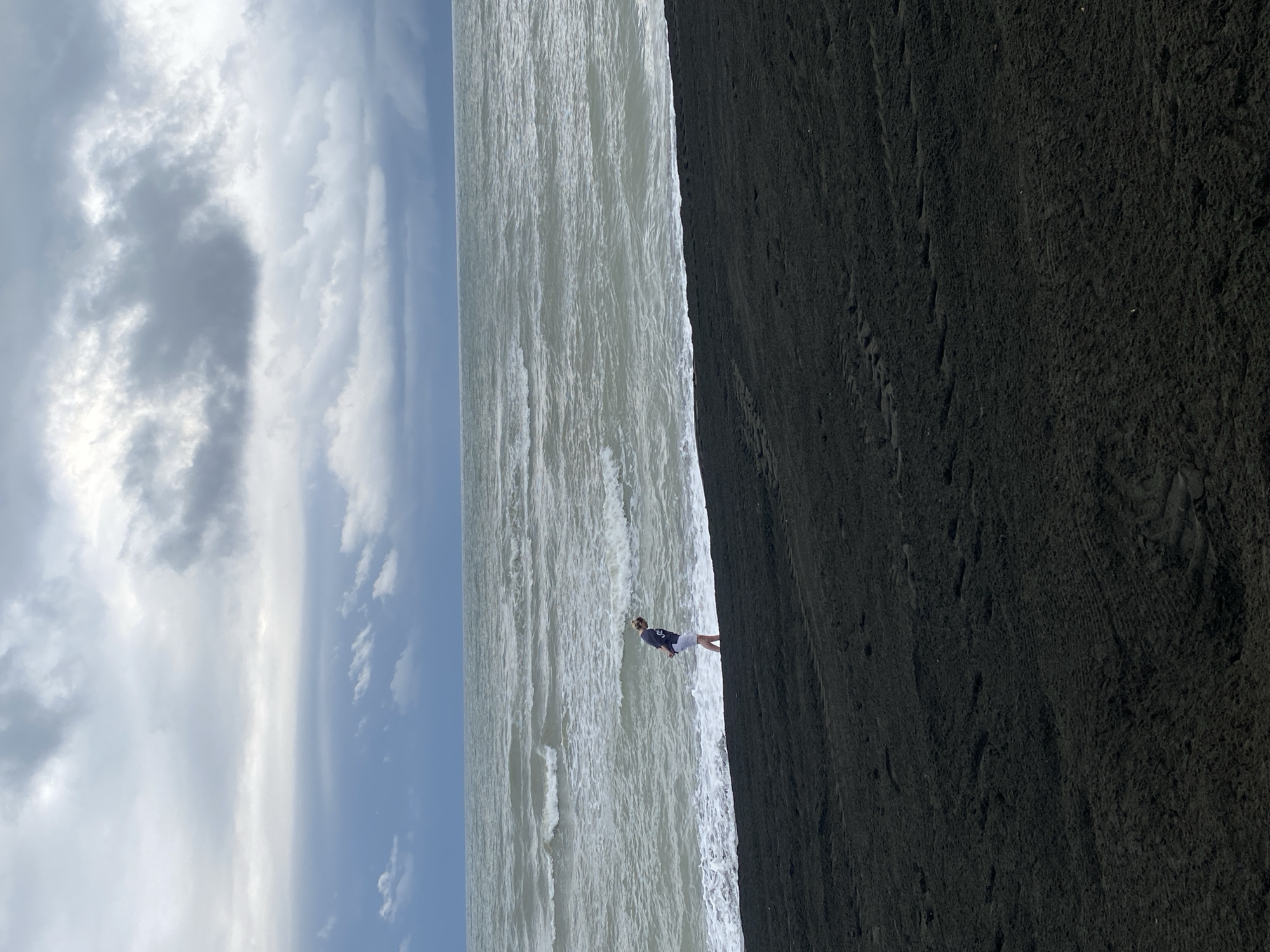A person walks along a black sand beach on a cloudy day. The person is wearing a dark blue t-shirt and white shorts, and their hands are behind their back. They are facing the ocean, which has waves breaking on the shore. The sky is filled with dark, ominous clouds. The beach itself is composed of fine black sand, with some tire tracks visible in the foreground. The location is the black sand beach in Ureki, Georgia, known for its therapeutic magnetic sand.