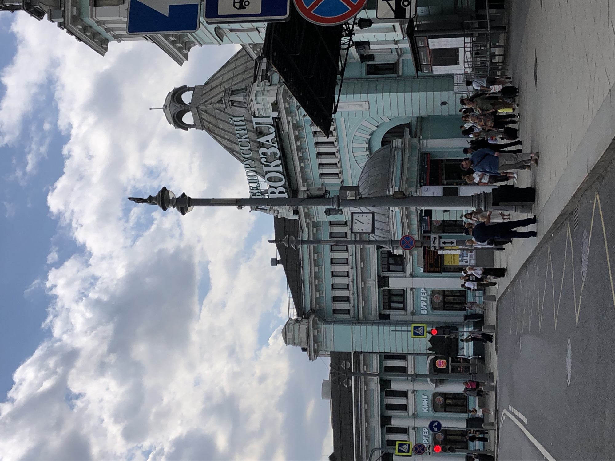 The image shows the Belorussky railway station in Moscow, Russia. It is a bright, cloudy day. The station is a large, light blue building with a prominent dome. People are walking on the sidewalk in front of the station, some with luggage. There are also several signs and traffic lights visible. A Burger King restaurant is located on the ground floor of the station. The street in front of the station is paved and there is a crosswalk. The overall scene depicts a busy urban environment with a focus on the historical architecture of the train station.