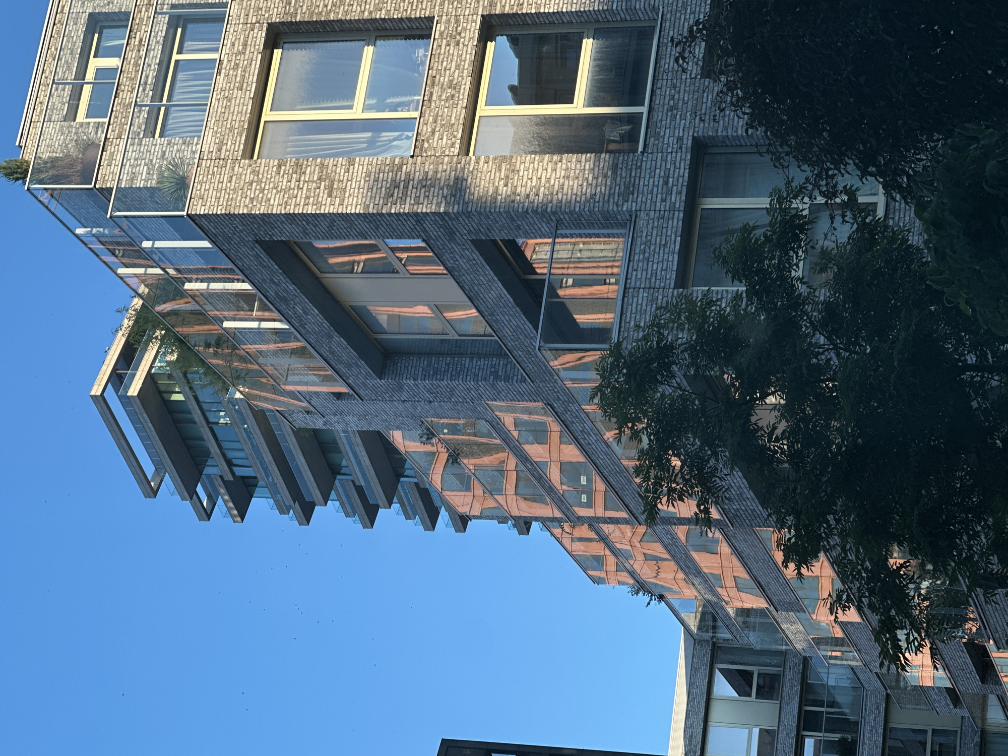 A low-angle, full shot captures a modern, multi-story building against a clear blue sky in Amsterdam, Netherlands. The building features a stepped design with numerous balconies and large windows. The facade is constructed with light gray bricks and accented with metal and glass. The sunlight, coming from the side, creates strong reflections on the windows of the neighboring building, displaying an orange hue. Green trees are visible in the foreground, partially obscuring the lower portion of the building. Several small, dark specks, possibly birds or insects, are scattered across the sky. The image appears to be taken during daylight hours, likely late afternoon or early evening due to the warm light.