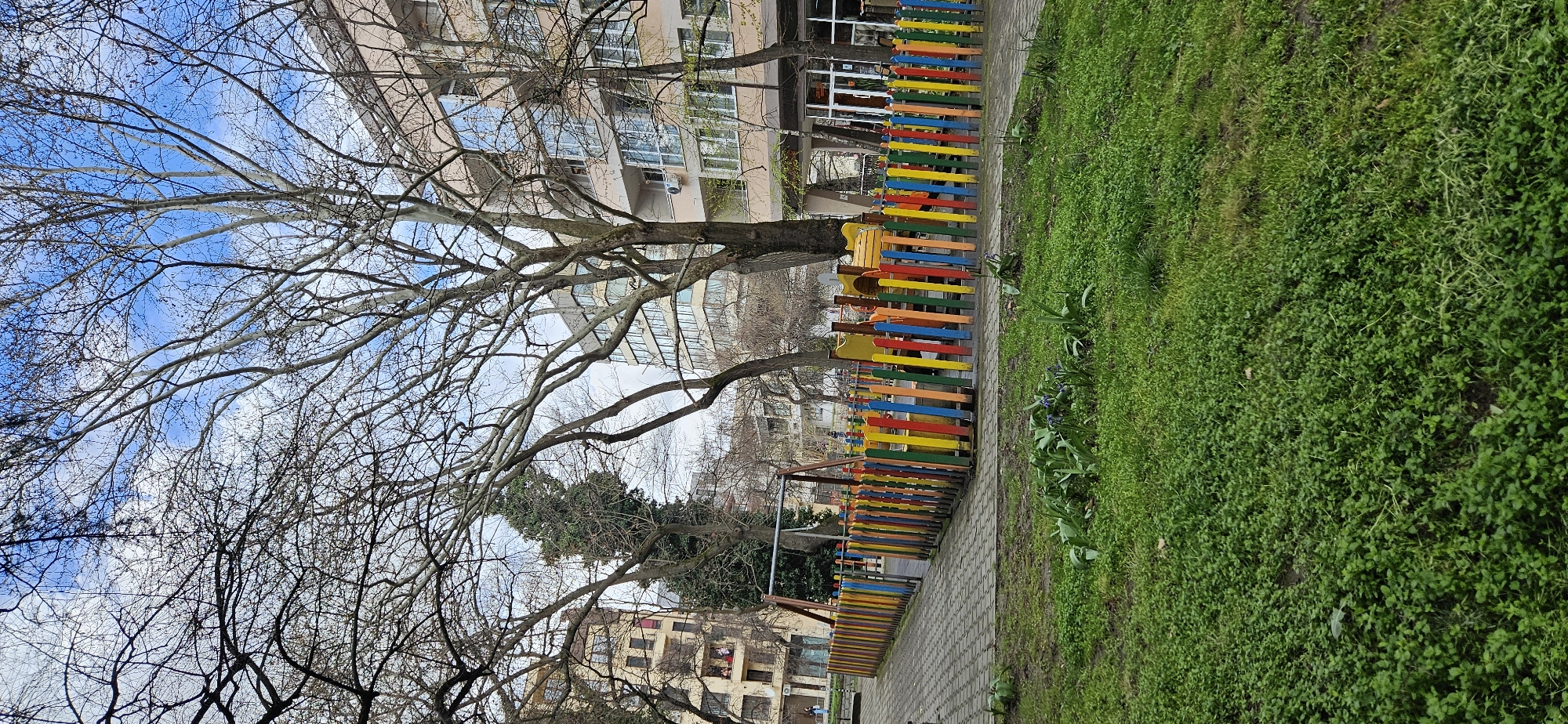 The image depicts an urban park scene in Blagoevgrad, Bulgaria. The park features a colorful wooden fence composed of horizontal slats in various colors (yellow, red, blue, green, orange). The fence borders a paved walkway and a patch of green grass. Mature trees are present, some with bare branches silhouetted against a sky with scattered clouds. Buildings are visible in the background, indicating an urban setting. No people are visible in the image.