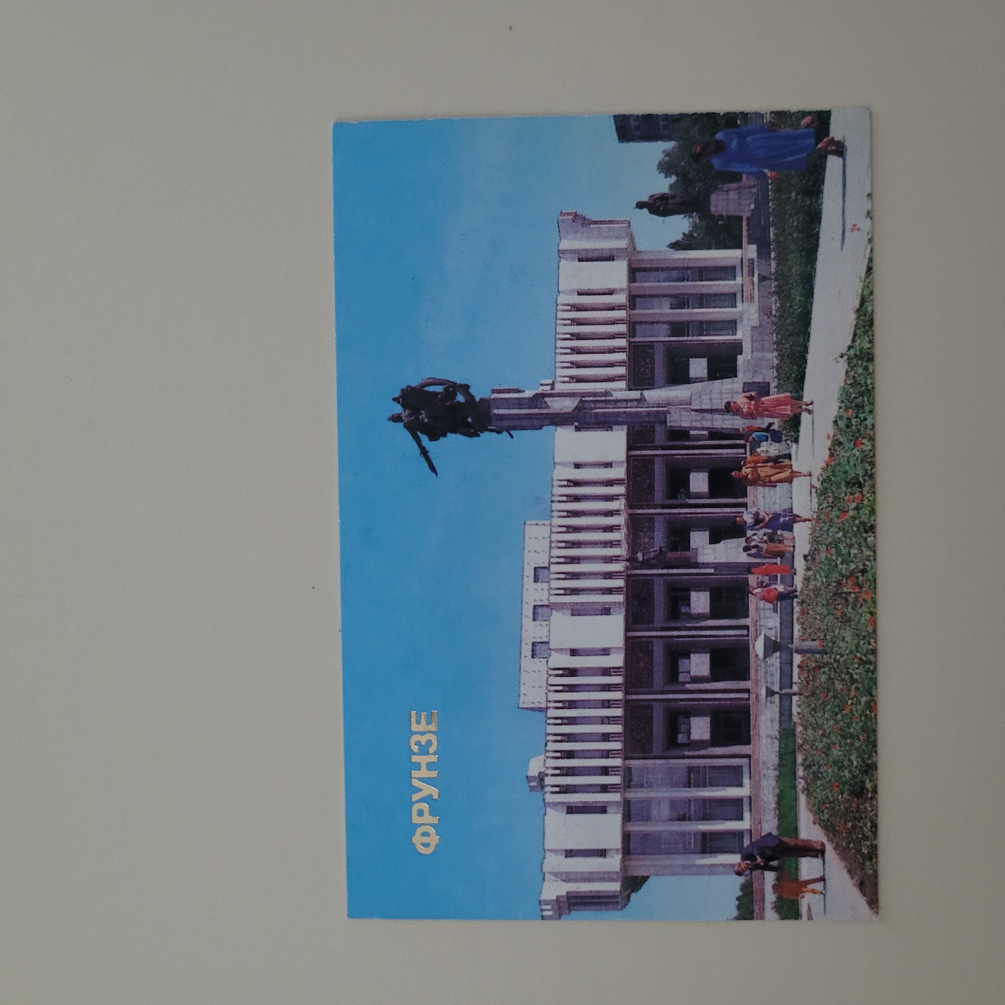 The image is a postcard with a light gray border. The postcard itself features a bright blue sky over a building and a monument. In the upper left quadrant of the postcard, the word "ФРУНЗЕ" is printed in a metallic gold font.
The building in the background is modern, with a white facade and numerous vertical columns. It has large, dark windows and a row of smaller square windows on the upper level.
In front of the building, a tall, dark monument stands. It is topped by a sculpture of two figures on horseback, appearing to be engaged in battle or movement. The monument itself is abstract and angular.
In the foreground, a paved walkway runs in front of the building and past a garden area with red flowers. Several people are depicted:
* On the left side of the walkway, a man in a dark suit and brown pants bends down towards a small child in an orange outfit.
* Further along the walkway, several people, mostly women in dresses, are walking. One woman in a yellow dress is prominently featured.
* On the right side of the walkway, a woman in a long blue dress is walking away from the camera.
* In the distance, to the right of the main building, another, smaller building can be seen.
* Behind the main building and to the right of the monument, a dark silhouette of a statue is visible.
The scene appears to be a public space, possibly a plaza or park area in front of a significant building. The image quality suggests it is likely an older photograph, possibly from the Soviet era, given the Cyrillic script and the style of the monument and building.
The location is the city of Jūrmala, Latvia.