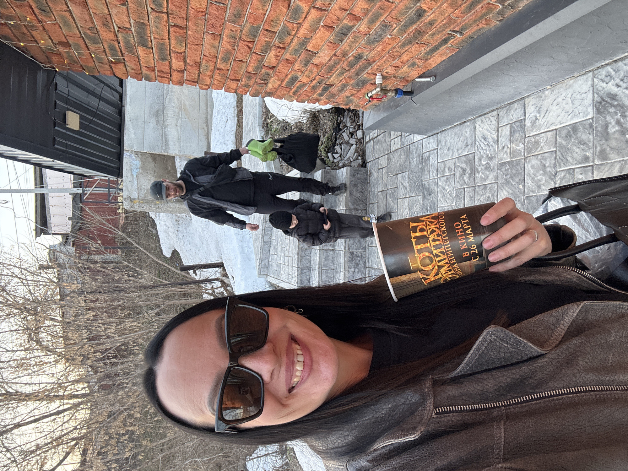 A selfie of a smiling woman wearing sunglasses and a leather jacket, holding a paper cup with the words "КОТЫ ЭРМИТАЖА В КИНО С 26 МАРТА," meaning "Hermitage Cats in Cinemas from March 26th" in Russian. In the background are two figures, one adult and one child, walking up a flight of outdoor stairs. There is snow on the steps and adjacent to the stairs. A brick wall is visible in the background, along with a section of a black structure. The scene appears to be outdoors in a residential or urban environment. Given the writing on the cup and the location of Chaikovsky, Russia, the image likely depicts the woman promoting a film about cats living in the Hermitage Museum.