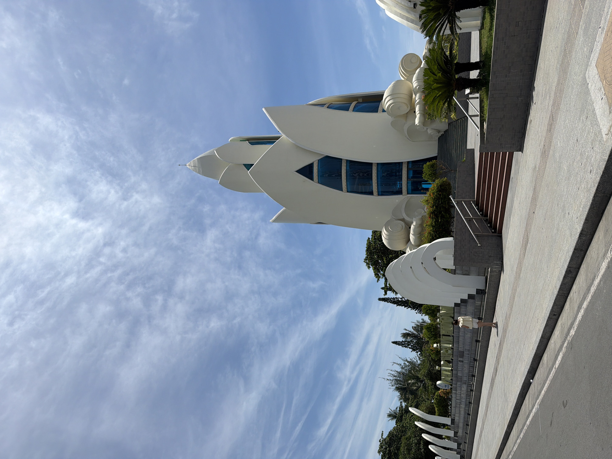 In Nha Trang Ward, Vietnam, a striking white building with a unique lotus-like design stands against a backdrop of a bright blue sky dotted with wispy clouds. The building features large, blue-tinted windows and decorative, swirling white elements at its base. In front of the building, a person wearing a light-colored outfit stands on a paved walkway, facing away from the camera. The walkway leads up a set of red-stepped stairs towards the building. To the left of the walkway, a series of curved white sculptures are arranged in a line, and beyond them, lush green trees and palm trees are visible. A paved road runs along the bottom of the image.