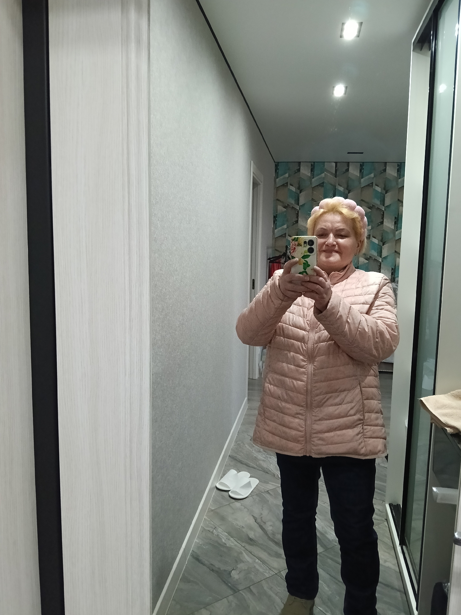 An elderly woman is taking a selfie in what appears to be a hallway. She's wearing a pink puffer jacket, dark pants, and a pink headband. She is holding a smartphone with a floral case. White slippers are on the floor behind her. The hallway has light grey walls and a tiled floor. Through an open doorway, wallpaper with a geometric pattern is visible. The location is likely in Murom, Russia, but it is based on the user's information.