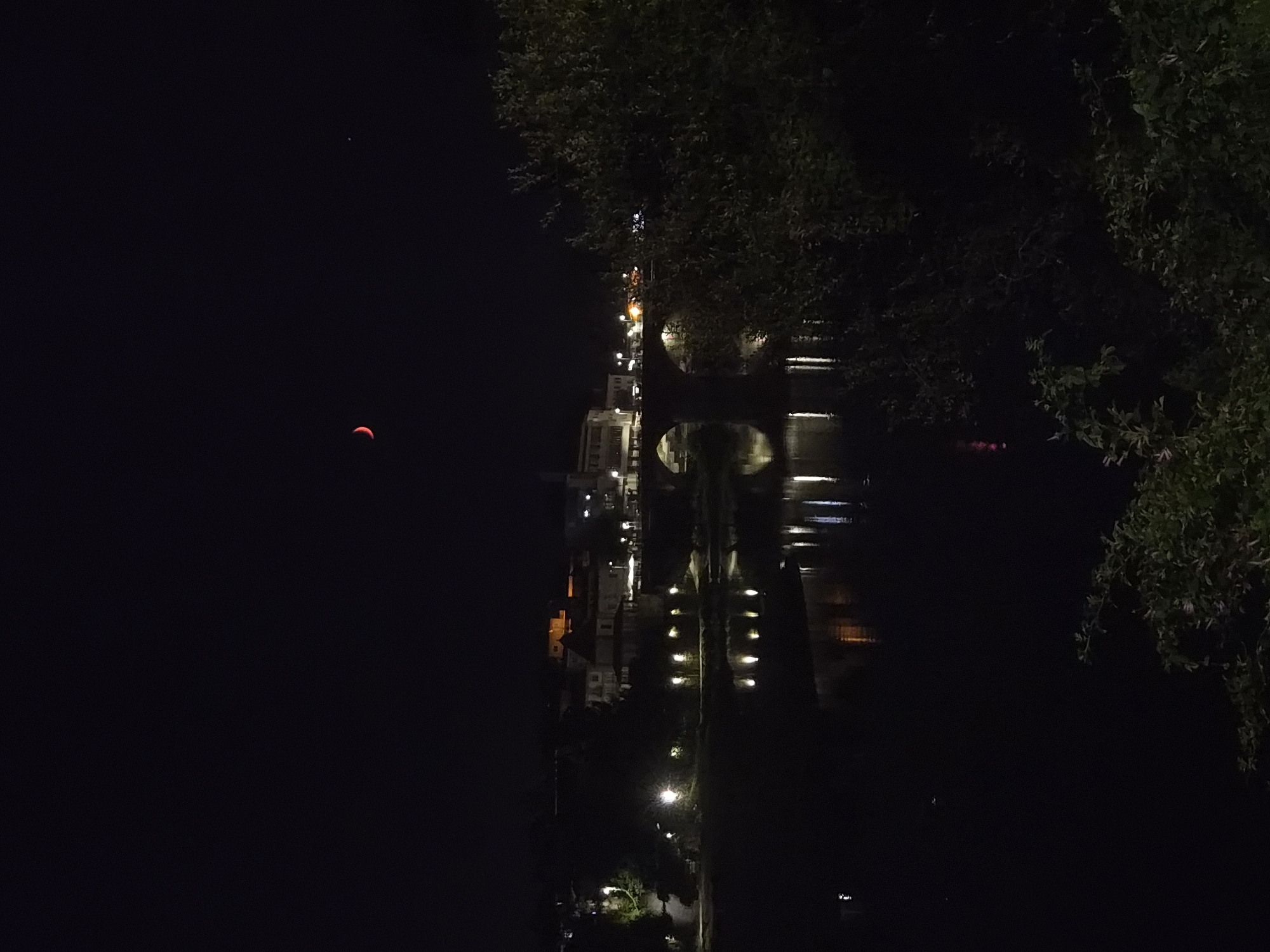 The image shows a nocturnal view of the city of Barcelos, Portugal, with a crescent moon and a distant star visible in the dark sky. A river flows in the foreground, reflecting the lights from the buildings and a bridge that spans the river. The bridge is illuminated by streetlights, and there are several buildings visible along the riverbank, some of which are lit up. Trees and vegetation are present on both sides of the river, adding to the natural landscape. The overall atmosphere is serene and tranquil, with the city lights creating a warm glow against the dark night.
