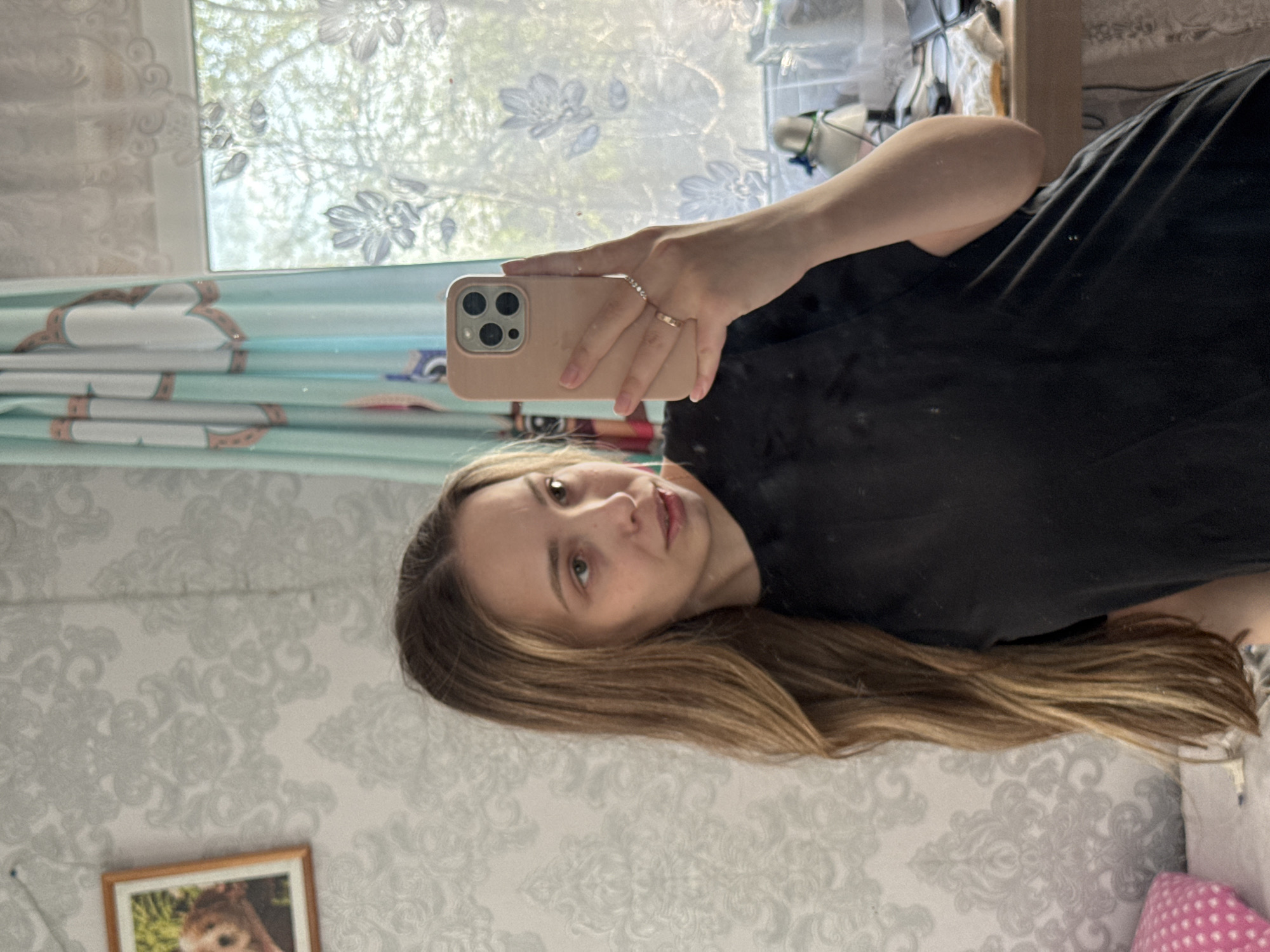 A woman is taking a selfie in what appears to be a room. She is holding a tan iPhone in her right hand and wearing two rings on that hand. She is wearing a black shirt. Her hair is blonde with darker roots. The room has patterned wallpaper and a window with blue floral curtains. In the window’s reflection, there are tree branches. A framed picture is on the wall near the bottom of the image. There is no explicit evidence that the scene is in the city of Кемуль, Russia.