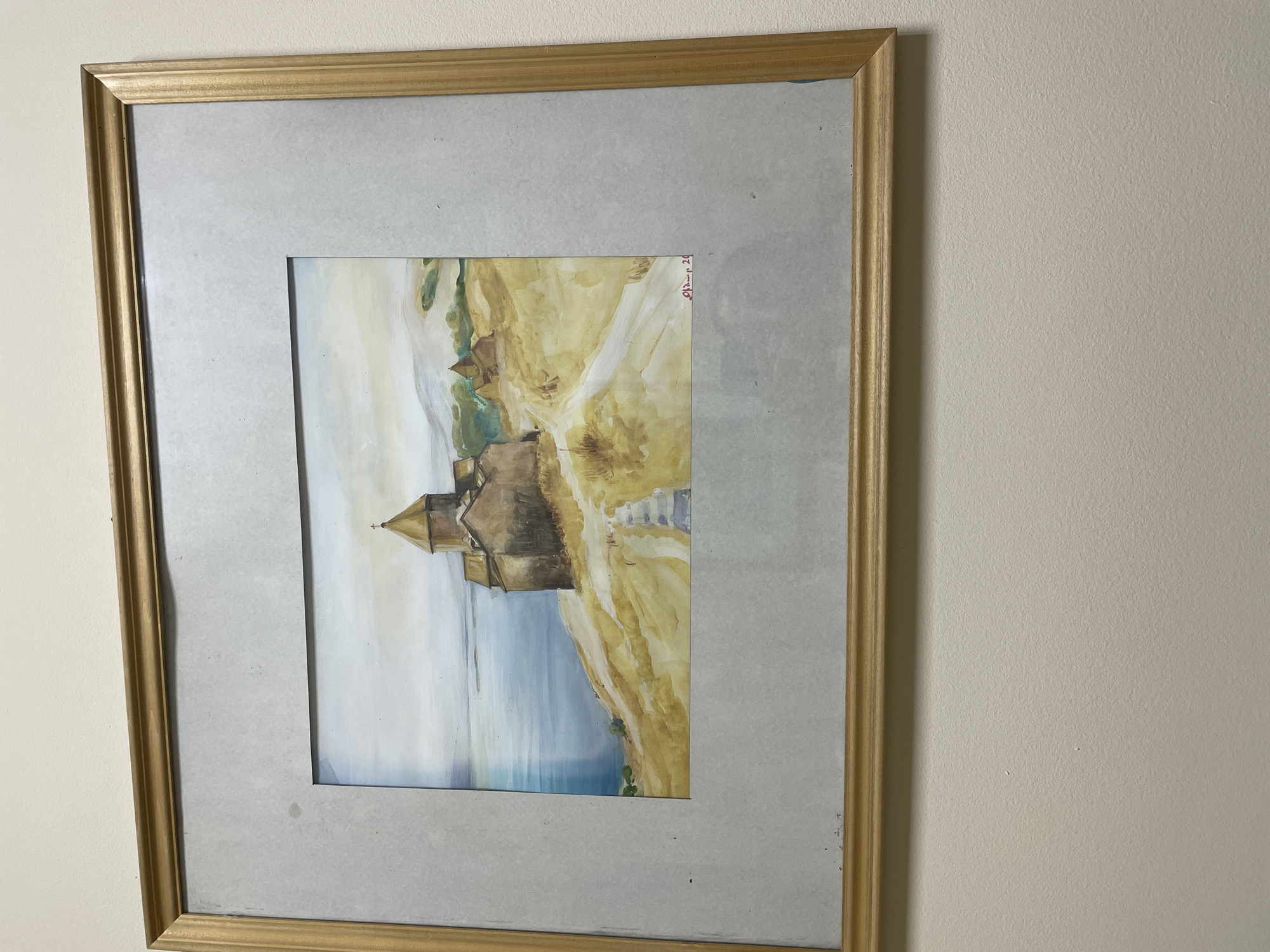 A rectangular framed watercolor painting hangs on a light beige, textured wall. The frame is golden-brown with a ridged profile, encasing a light gray matboard with minor dust specks. The painting depicts Sevanavank Monastery, an Armenian monastic complex, situated prominently on a golden-brown, arid-looking hill or peninsula. The monastery features several stone structures, including a main building with a distinct polygonal dome topped by a small cross. A light-colored path ascends the hill towards the monastery. To the left, a calm blue body of water, identifiable as Lake Sevan, stretches towards the horizon under a pale, overcast sky. Smaller, simpler brown buildings are visible behind the main monastery structure, and faint green hills appear in the distant background. A red signature or inscription, reading "Թաշչյան 20" or similar, is located in the lower right corner of the artwork. Sevanavank Monastery is located on a peninsula on Lake Sevan in Gegharkunik Province, Armenia, not within the city of Yerevan.