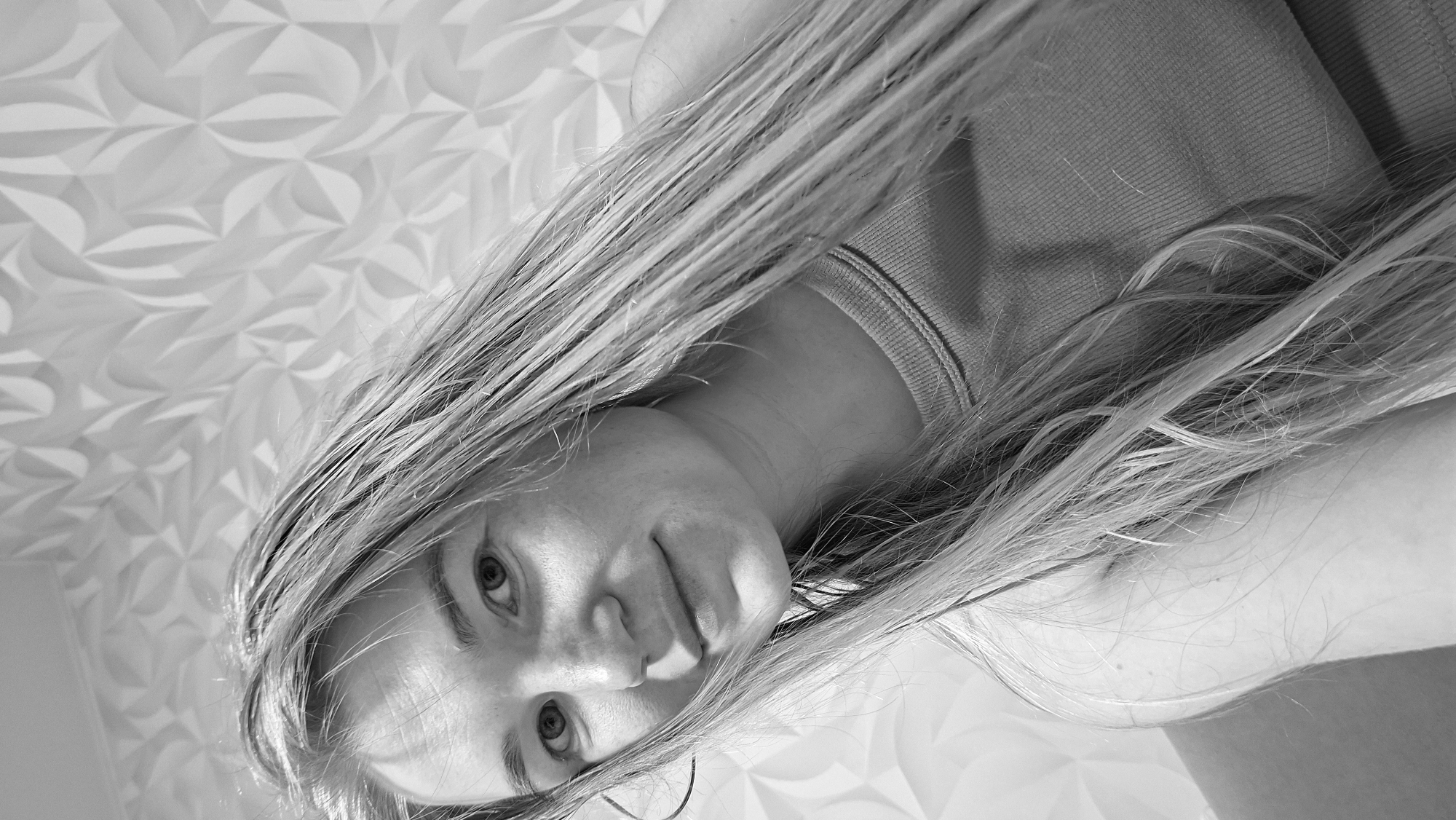 A black and white, waist-up, medium shot shows a young woman with long, blonde hair, possibly wet, that falls around her shoulders. She is looking at the camera with a slight smile, her head tilted to her left. Her eyes are open, and she appears to be wearing a simple, sleeveless top. The background is a textured wall with a geometric pattern. The overall lighting suggests an indoor setting. The image does not provide any specific indicators of its location within the city of Chaikovsky, Russia.