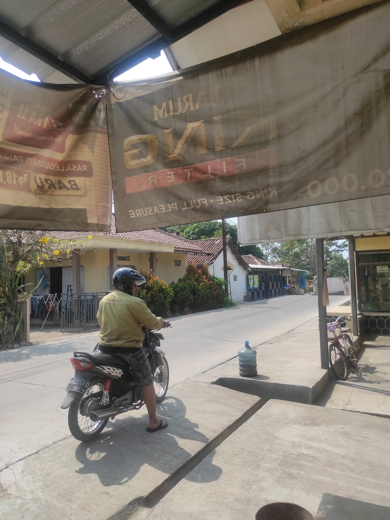 A person wearing a yellow jacket and dark camouflage shorts is seated on a black motorcycle, facing away from the camera, on a concrete sidewalk. They are wearing a black helmet and dark flip-flops. The motorcycle has a license plate that reads "5822 FC". To the left of the motorcycle, there is a fence and laundry hanging on a clothesline in front of a light yellow house with a red-tiled roof. A tree with yellow flowers is also visible near the house. Across the street, more houses with red-tiled roofs and trees are visible. A large blue water jug is placed on the sidewalk to the right of the motorcycle. Further to the right, a purple bicycle with a basket is leaning against a pole. Above the scene, a large, faded banner is stretched across, displaying text in Indonesian. The sky is bright and sunny, casting shadows on the ground. The overall environment suggests a residential or commercial area in a town or city. This scene is located in Ngamprah, Indonesia, possibly on a local street.