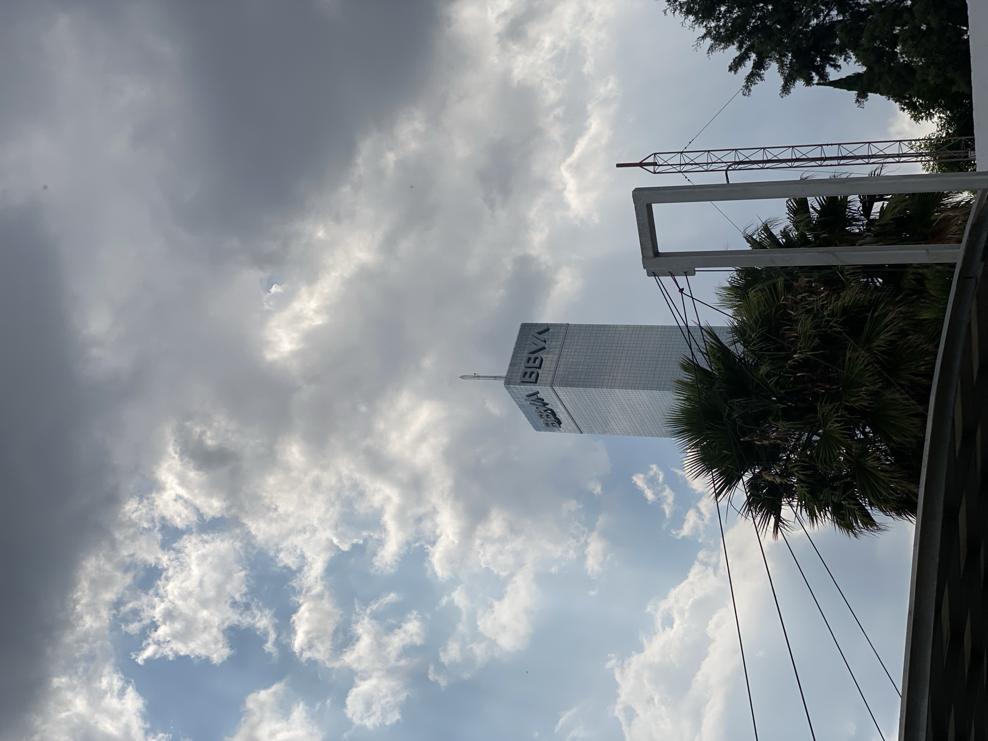 The image shows a tall building with the word "BBVA" on its side, partially obscured by clouds. The sky is filled with a mix of dark, heavy clouds and patches of blue sky. In the foreground, there's a palm tree and part of a metal structure, possibly a crane or framework. The location appears to be urban, based on the skyscraper and surrounding structures. The image suggests a cloudy day in an urban area.