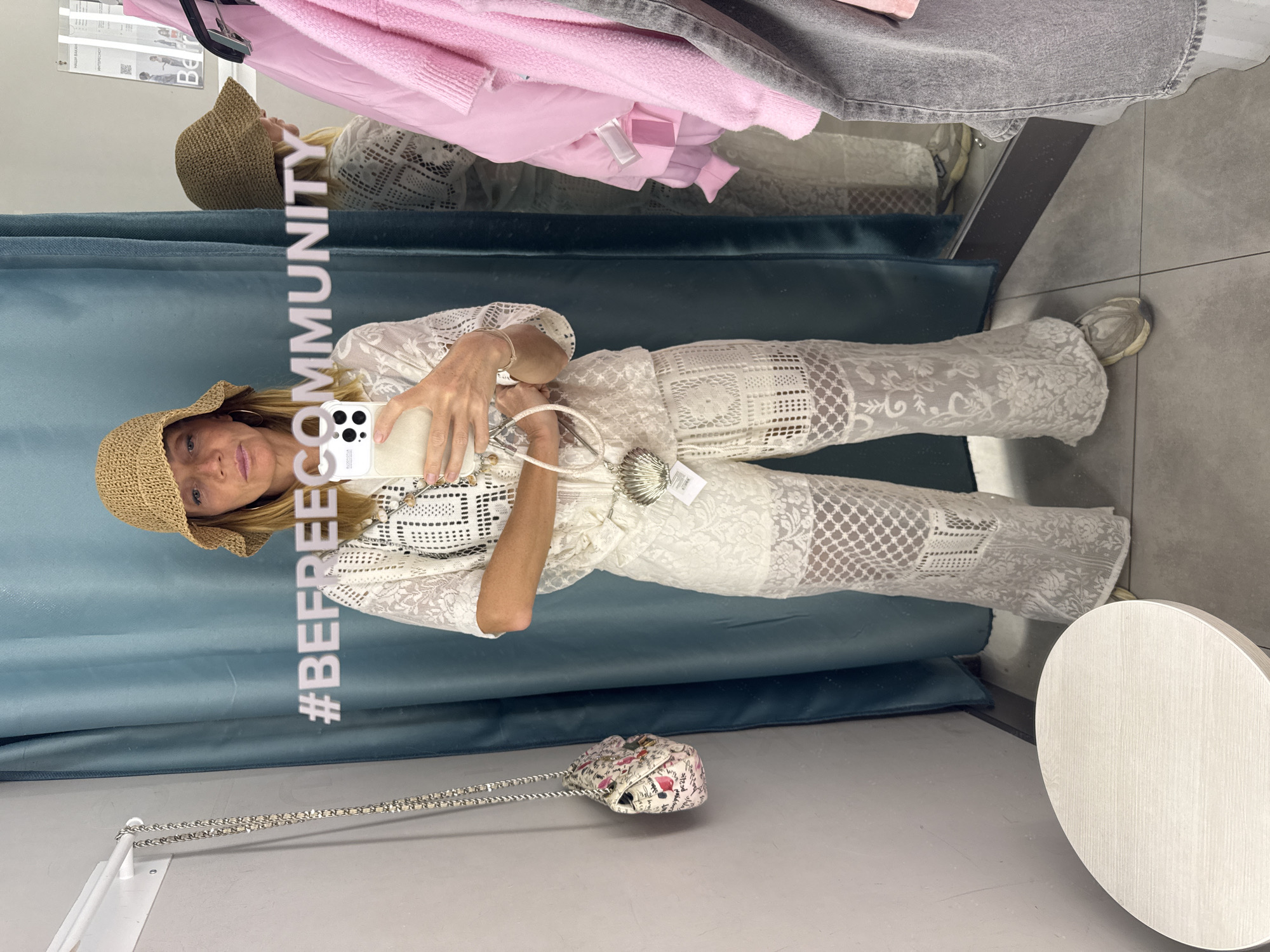 A woman is taking a mirror selfie in a fitting room. She is wearing a white lace outfit and a straw hat. She is holding a white smartphone in her hand. In the background, there is a blue curtain and a round white table. There are clothes hanging above the mirror. The hashtag "#BEFREECOMMUNITY" is visible on the mirror. The location is likely in a clothing store, though specific details to pinpoint Saint Petersburg, Russia, are missing from the image.