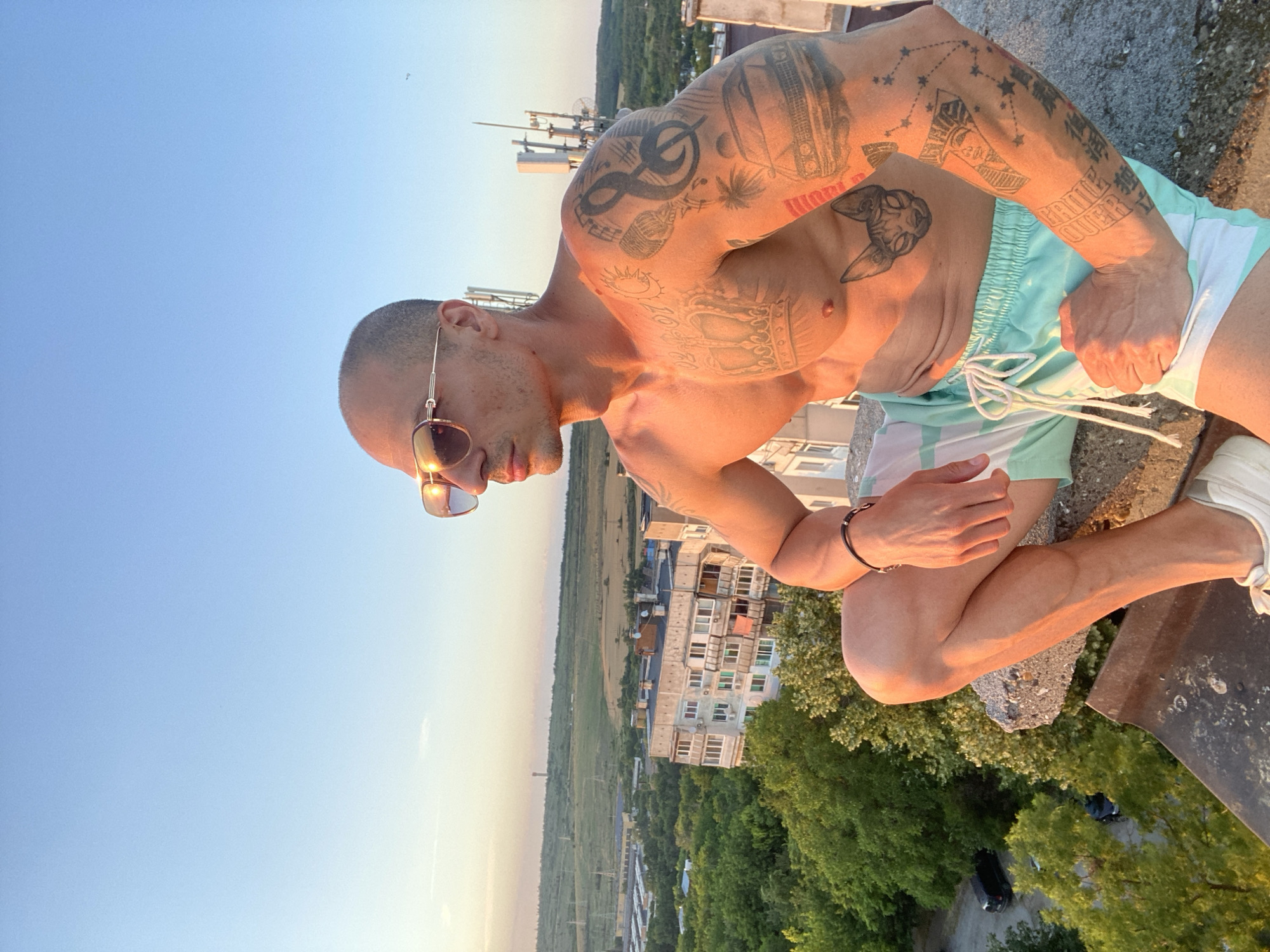 A bald man with sunglasses and tattoos is sitting on the edge of a building, looking out over a city. He is wearing swim trunks. The sun is setting, casting a warm glow on the scene. The city in the background appears to be Pleven, Bulgaria. The man has several tattoos, including a crown on his chest, a cat on his torso, and various designs on his arm. The image is a selfie taken from a high angle.