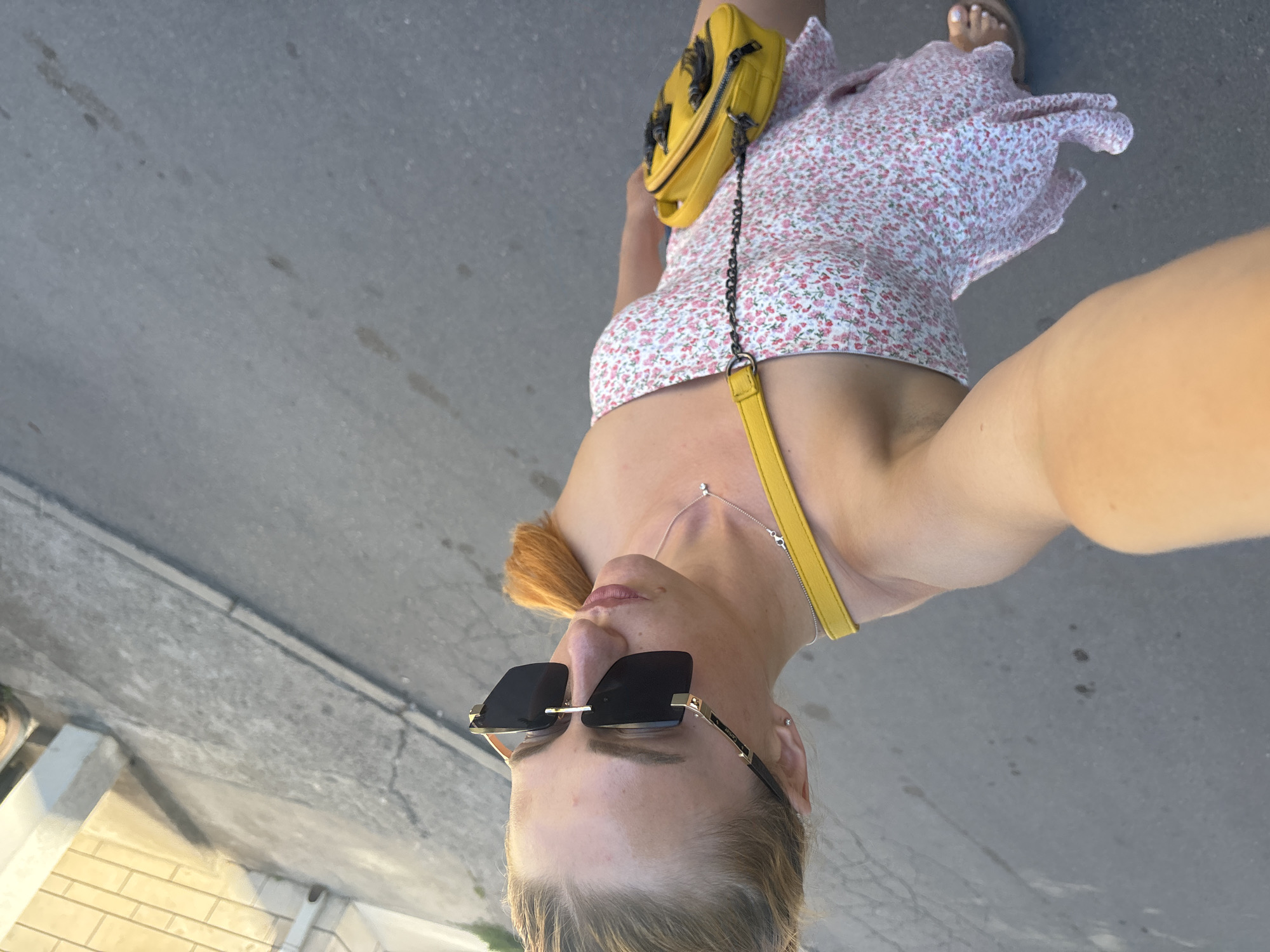 A woman is taking a selfie while standing outdoors on a paved surface, likely a sidewalk or street. She is wearing a short, floral print dress with a pink and white pattern and a yellow crossbody bag with a chain strap. She also has on large, black-framed sunglasses and a delicate necklace. Her hair is light blonde and pulled back. The photo appears to be taken during the daytime, as indicated by the bright lighting and shadows. The setting seems to be an urban or semi-urban environment, with the pavement and what looks like a building or curb in the background. The image is oriented vertically, and the woman's face is positioned at the bottom. The overall mood is casual and sunny. No visible text is present.