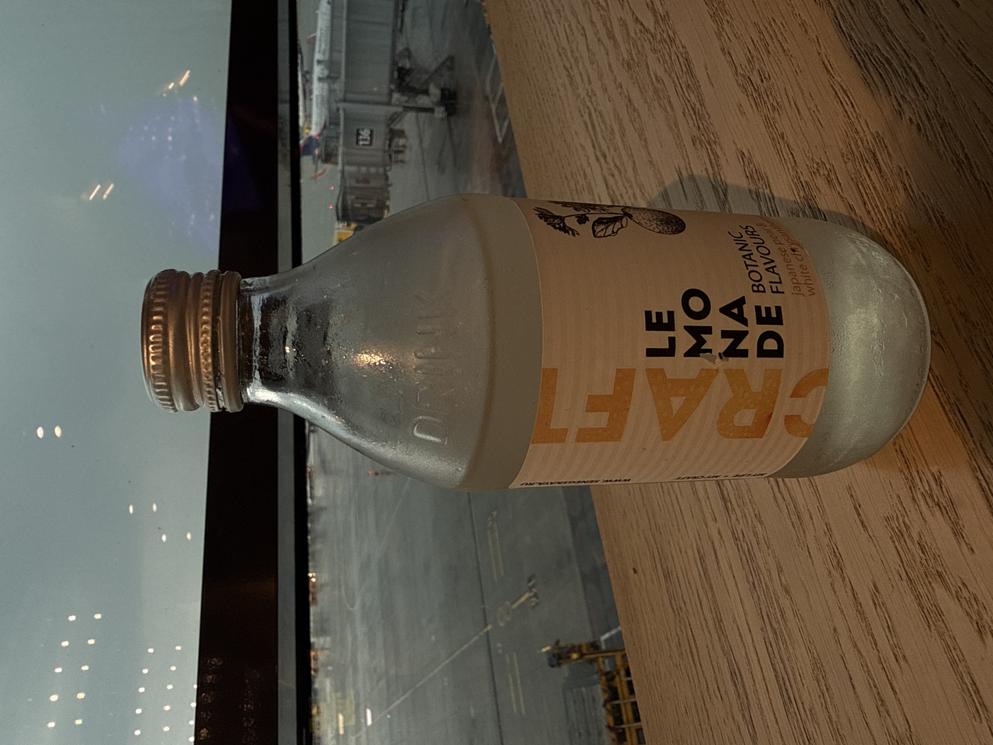 A close-up, eye-level, medium shot captures a glass bottle of "CRAFT LEMONADE" with "BOTANIC FLAVOURS" of "japanese pear & white chrysanthemum" on a wooden surface, likely a table. The bottle is condensation-covered. In the background, a large window overlooks an airport tarmac at dusk or dawn. Visible are an airport terminal with a jet bridge numbered "136," an airplane, and runway lights. The sky is overcast, with reflections of interior lights visible. The overall atmosphere suggests a travel waiting area, possibly at Sheremetyevo International Airport, located in Khimki, Russia.