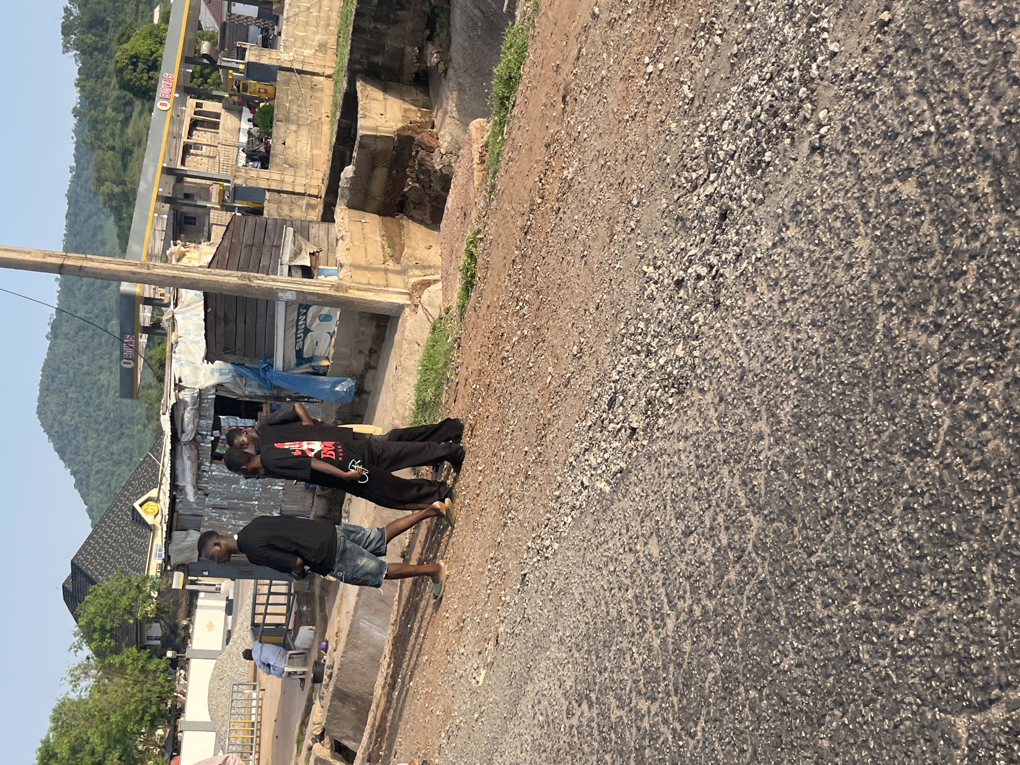 A group of four young men are walking on a gravelly road in front of a gas station and some small buildings. The men are dressed casually. One man is wearing a black t-shirt and denim shorts, another is wearing a black t-shirt with a graphic design, and two are wearing black t-shirts and black pants. In the background, there is a Bovas gas station with a yellow and gray awning, and several small structures made of corrugated metal and concrete blocks. A person is sitting on a chair in front of a white building to the left. The area appears to be a rural or semi-urban environment, with a hill covered in trees in the distance. The sun is casting long shadows, suggesting it is late afternoon.