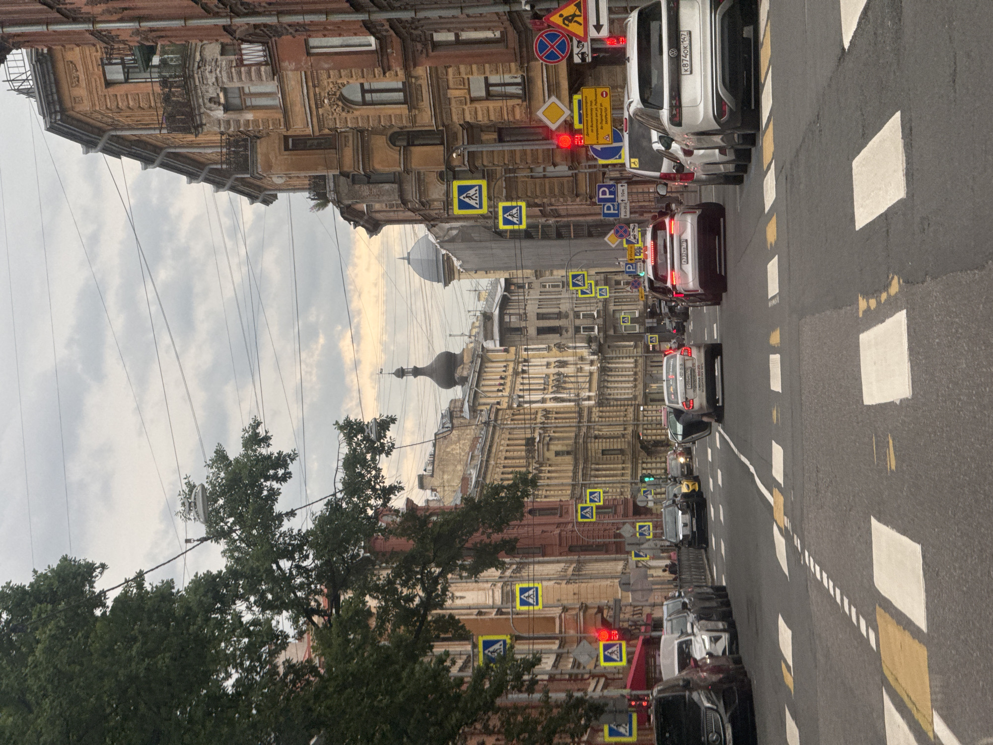 The image depicts a street scene in Saint Petersburg, Russia, likely during the daytime given the lighting. The perspective is from a vehicle, looking forward along a street lined with ornate, historic buildings. The architecture is characterized by detailed facades, balconies, and classical elements, suggesting an older part of the city.
The street is moderately busy with traffic. Several cars and a bus are visible, stopped or moving slowly, indicating a potential traffic jam or a busy intersection. There are numerous road signs, including pedestrian crossing signs (indicated by the "A" in a blue square), directional signs, and what appears to be a "No Entry" or "Closed" sign in Russian. Some signs have text that is partially legible, such as "Парадной ул" (Paradnoya Street) and "ЗАКРЫТО!" (CLOSED!). Other text includes vehicle license plates, for example, "K8740K 147" and "A732AB 778".
The sky is overcast with some patches of brighter sky, suggesting a cloudy or partly cloudy day. Trees with green foliage are visible on the left side of the image, adding a touch of nature to the urban environment. Overhead, a network of power or telecommunication lines crisscrosses the sky, a common sight in many cities. The road surface shows clear lane markings, including pedestrian crossing areas with white and yellow stripes. The overall impression is of a typical urban scene in a historic European city.