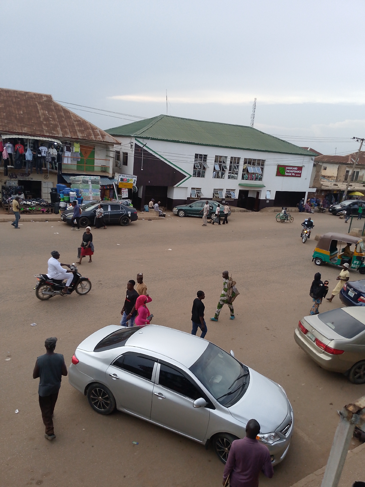 The image shows a street scene in Bukuru, Nigeria. The ground is unpaved and dusty. There are several people walking and standing around, some crossing the street. A man in white is riding a motorcycle. There are also several parked and moving vehicles, including cars, a motorcycle, and a three-wheeled auto-rickshaw (tuk-tuk). The building on the left has clothes hanging outside on display and what appears to be a storefront. Another building in the background has a sign that reads "HIGHLAND MICROFINANCE BANK Plc." The sky is overcast.