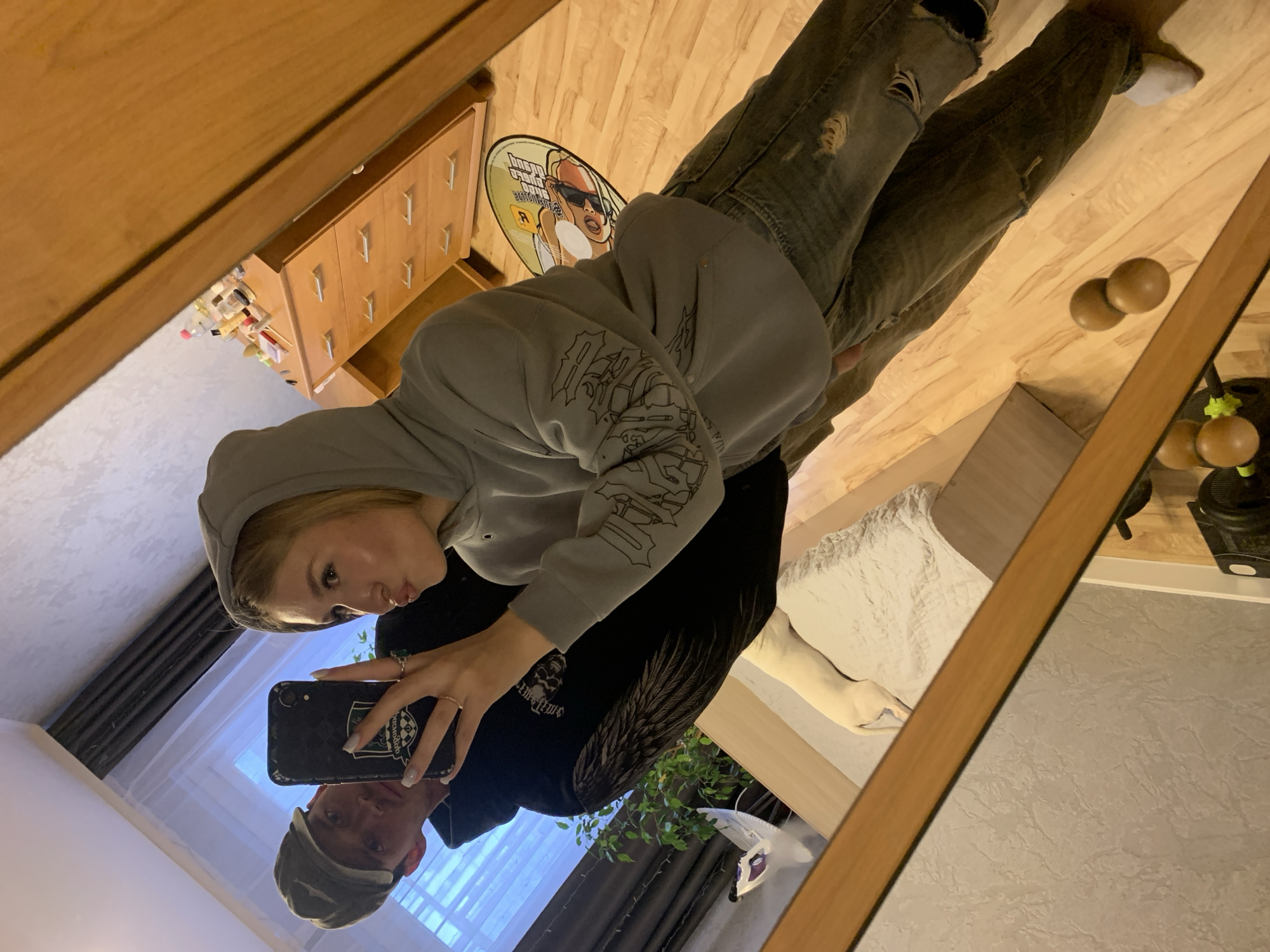 The image is a reflection in a mirror showing a room interior. Two people are discernible: a young woman holding a smartphone taking a selfie, and a man reflected below her. The woman wears a light gray hoodie with designs on the sleeve and a black garment underneath. The man is wearing a gray hat. The mirror shows their reflections in a room. Visible items include a wooden floor, a dresser with drawers, a GTA-themed floor mat, ripped jeans, a bed with white bedding, houseplants, and a floor lamp. A white dog lies on the bed. The lighting is soft, suggesting an indoor setting. The location of Chaikovsky, Russia is not discernable from the image.