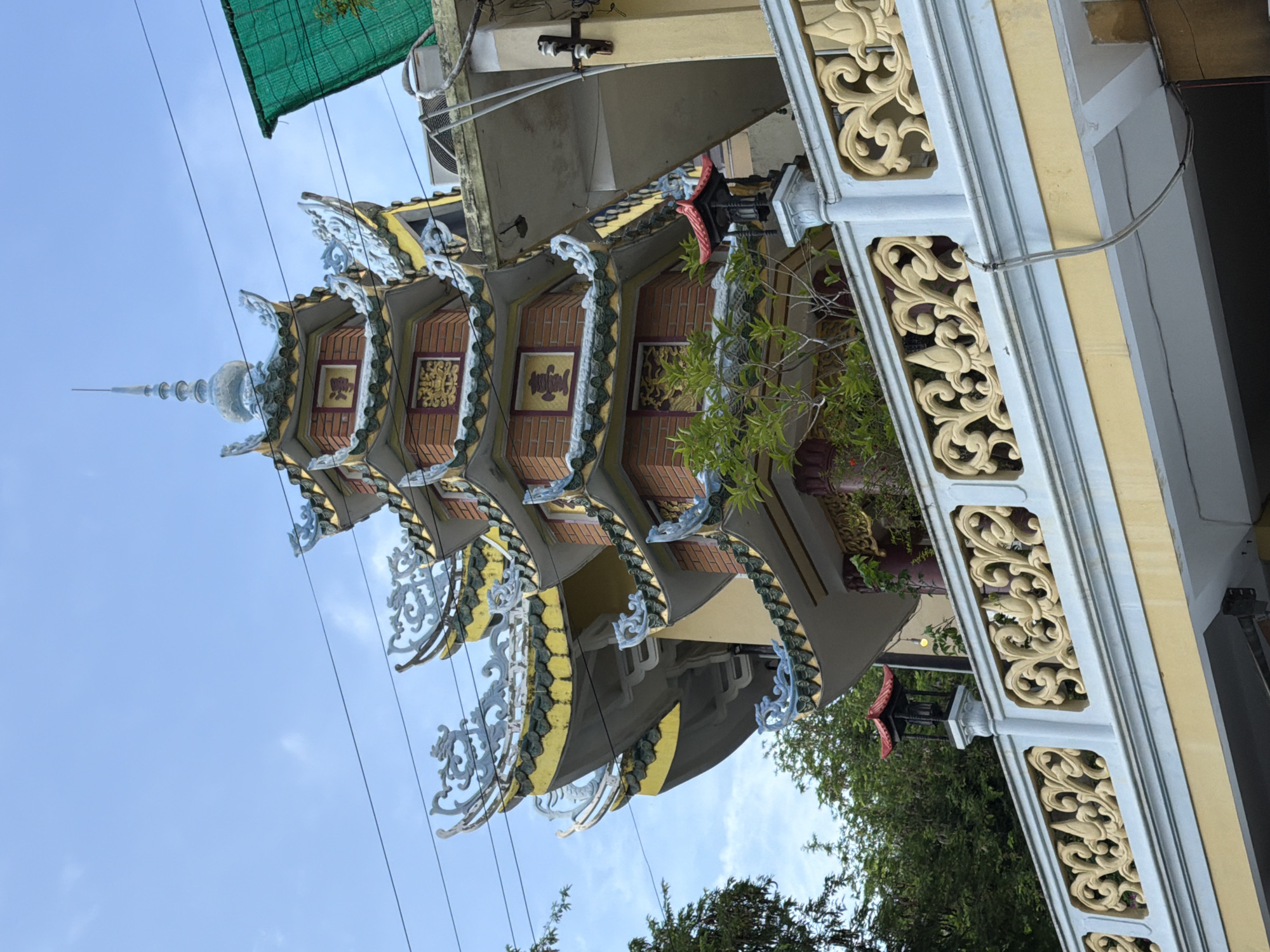 This is a close-up, eye-level, exterior, daytime, natural light shot of a multi-tiered pagoda with ornate, upturned eaves and dragon-like decorations. The pagoda is constructed with red brick and adorned with yellow and blue accents. It is situated behind a decorative light-colored railing with ornate carvings, and beyond that, a yellow wall and walkway. Lush green trees are visible behind and to the left of the pagoda. The sky above is a light blue with scattered white clouds. There are power lines running across the sky. A green tarp is visible on the upper right, partially obscuring a modern building structure. This pagoda is part of the Phat Tich Pagoda, located at 25 Tran Phu Street in Nha Trang, Vietnam. There are no people visible in the image.