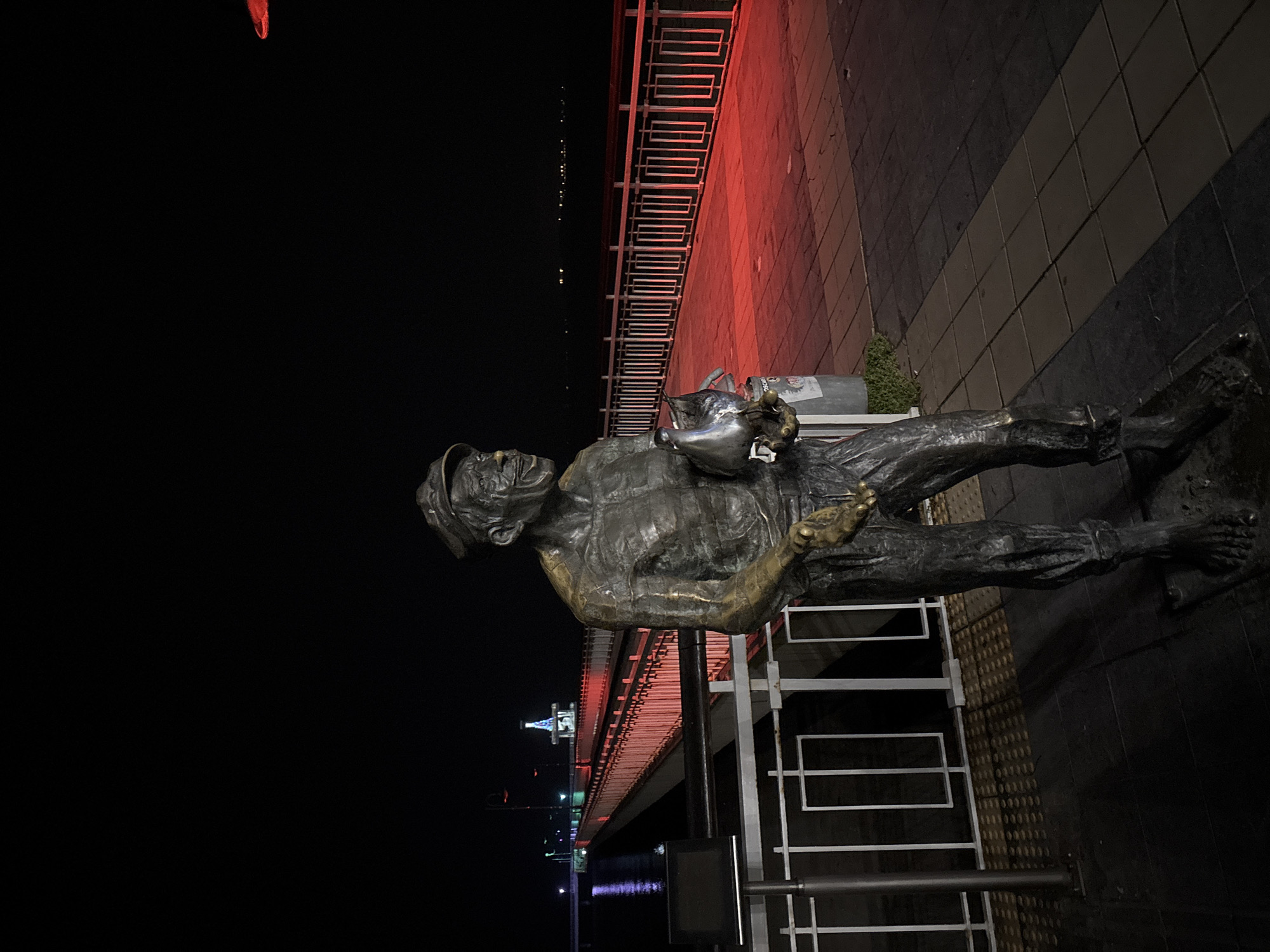 The image shows a bronze statue of a fisherman mending nets at night in Burgas, Bulgaria. The statue is positioned on a walkway near a bridge. The area is illuminated by red and blue lights reflecting off the water. The background is dark, suggesting it's nighttime. There is metal railing visible.