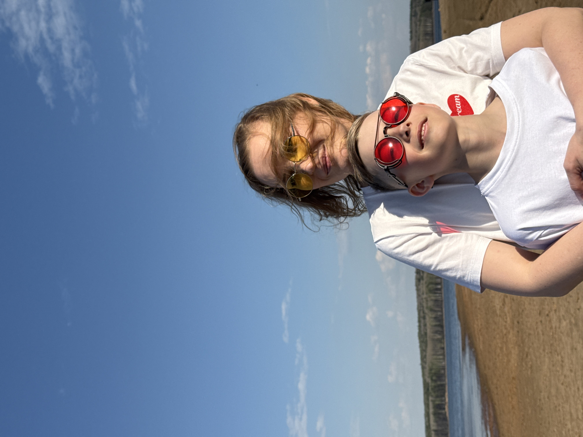 Two individuals are positioned near a body of water, possibly a lake or river, with a sandy shoreline visible. One person, wearing yellow sunglasses, is standing behind and slightly to the side of another person wearing red sunglasses. Both are wearing white t-shirts. The setting appears to be outdoors on a sunny day, with a clear blue sky and scattered clouds. Based on the prompt, the location is Chaikovsky, Russia. The scene appears to be a casual, candid moment captured outdoors.
