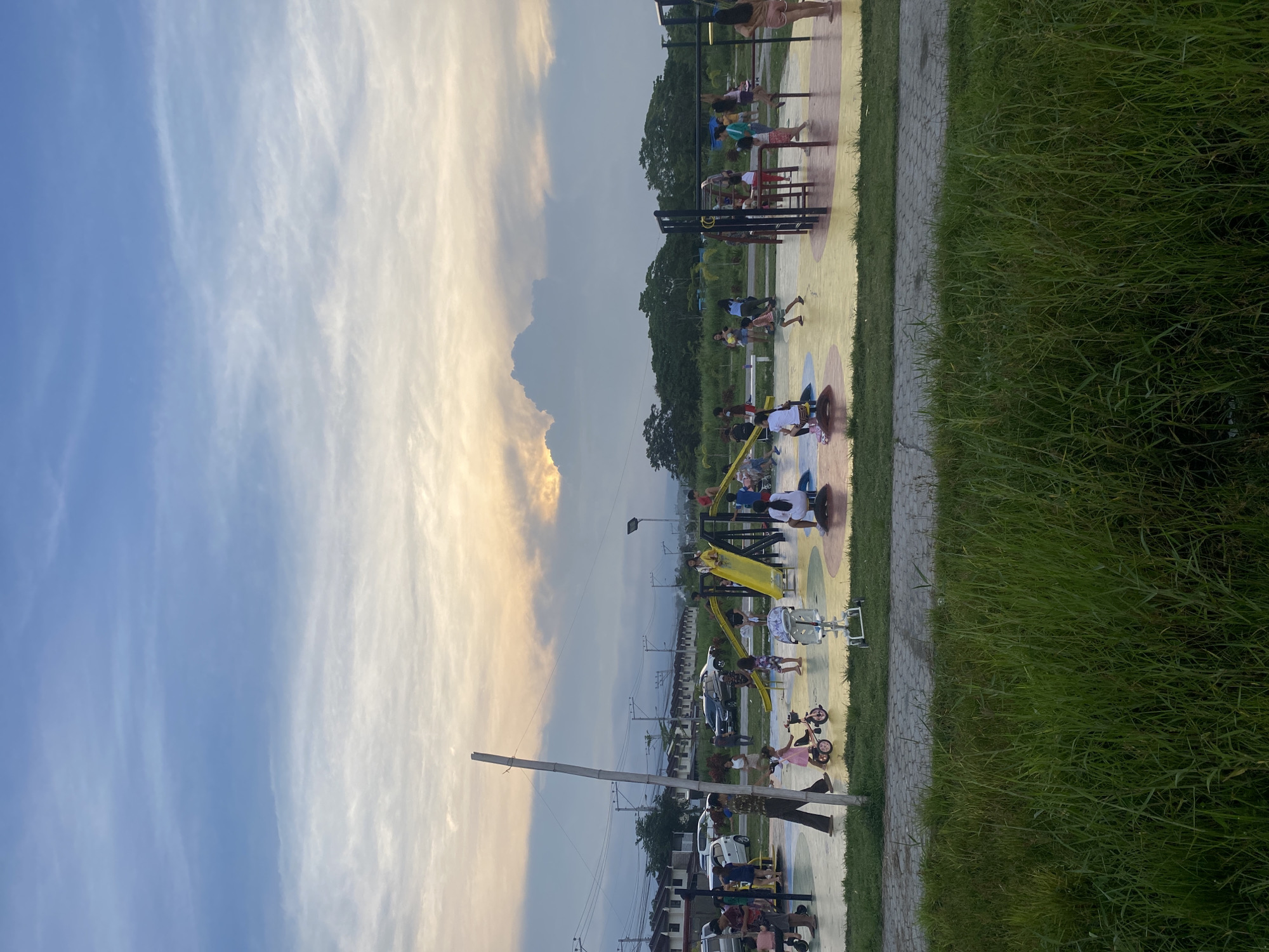 The image depicts a lively outdoor park or recreational area in Davao City, Philippines, during what appears to be late afternoon or early evening, characterized by a sky with a blend of clear blue and warm, golden-orange sunset hues filtered through wispy clouds.
The scene is bustling with people of various ages. In the midground and foreground, a colorful playground area with yellow, blue, and brown circular patterns on the ground features multiple pieces of equipment. Children are actively playing on yellow slides, monkey bars, and other structures. Adults are also present, some seemingly supervising children, while others are utilizing outdoor exercise equipment. A paved pathway separates the grassy area from the playground.
To the right, a lush strip of green grass borders the pathway. In the background, beyond the playground, are numerous trees, utility poles with a dense network of wires, and what appear to be distant residential buildings and parked vehicles. The overall atmosphere is one of leisure and community engagement, with people enjoying recreational activities. No visible text is discernable in the image.
