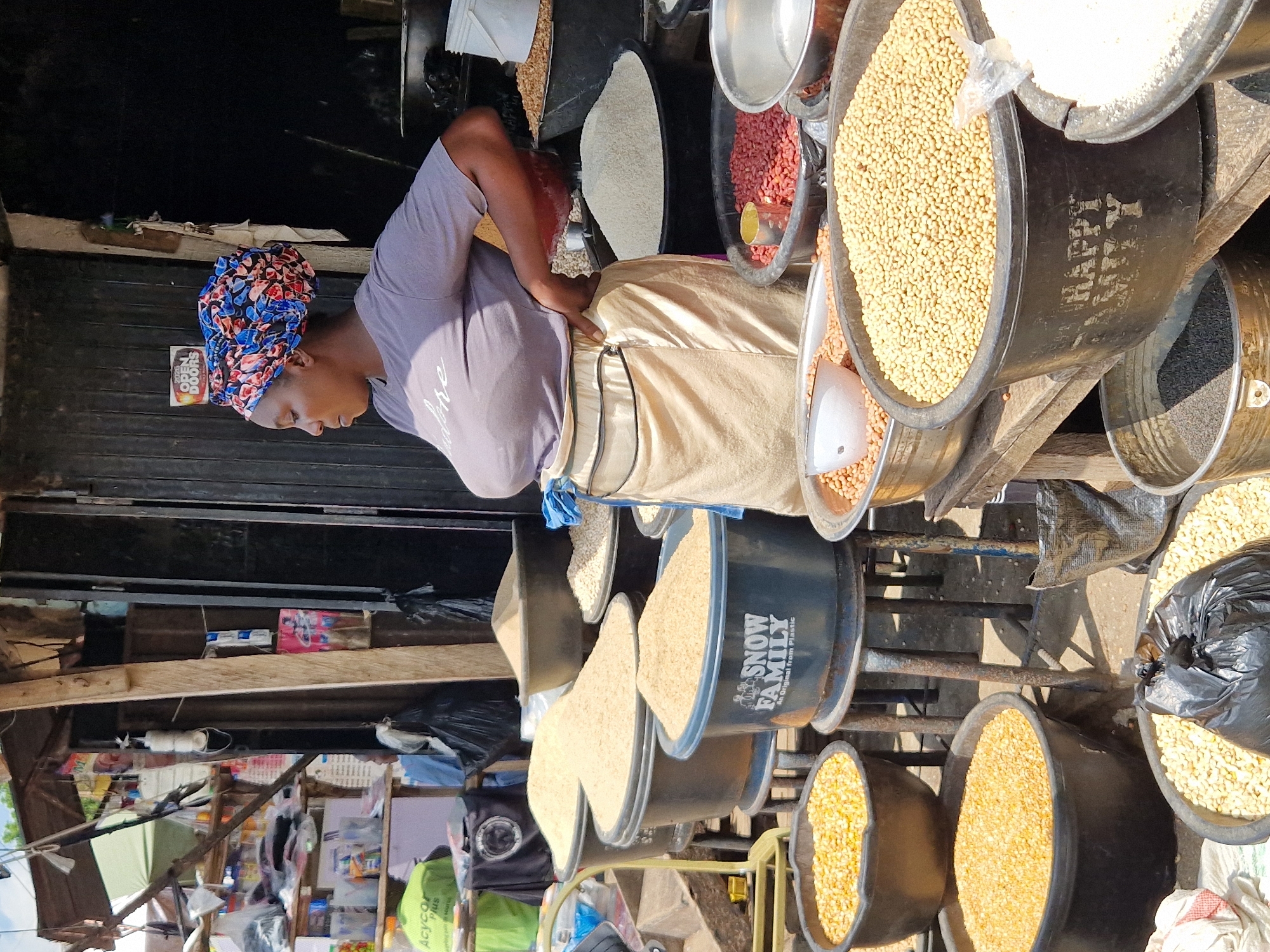 This image depicts a market scene in Jos, Nigeria. A woman wearing a colorful headwrap and a grey t-shirt stands amidst piles of grains and legumes displayed in large containers. She has her hand on her hip, and her gaze is directed to the right. Behind her, a stall is visible with various items on shelves, including what appear to be packaged goods. The ground is dusty, indicating an outdoor market setting. Several large black buckets are filled with different types of grains, such as what appears to be corn, rice, and beans. Some of these buckets have labels, including one that reads "SNOW FAMILY". The lighting suggests it is daytime, with sunlight casting shadows.