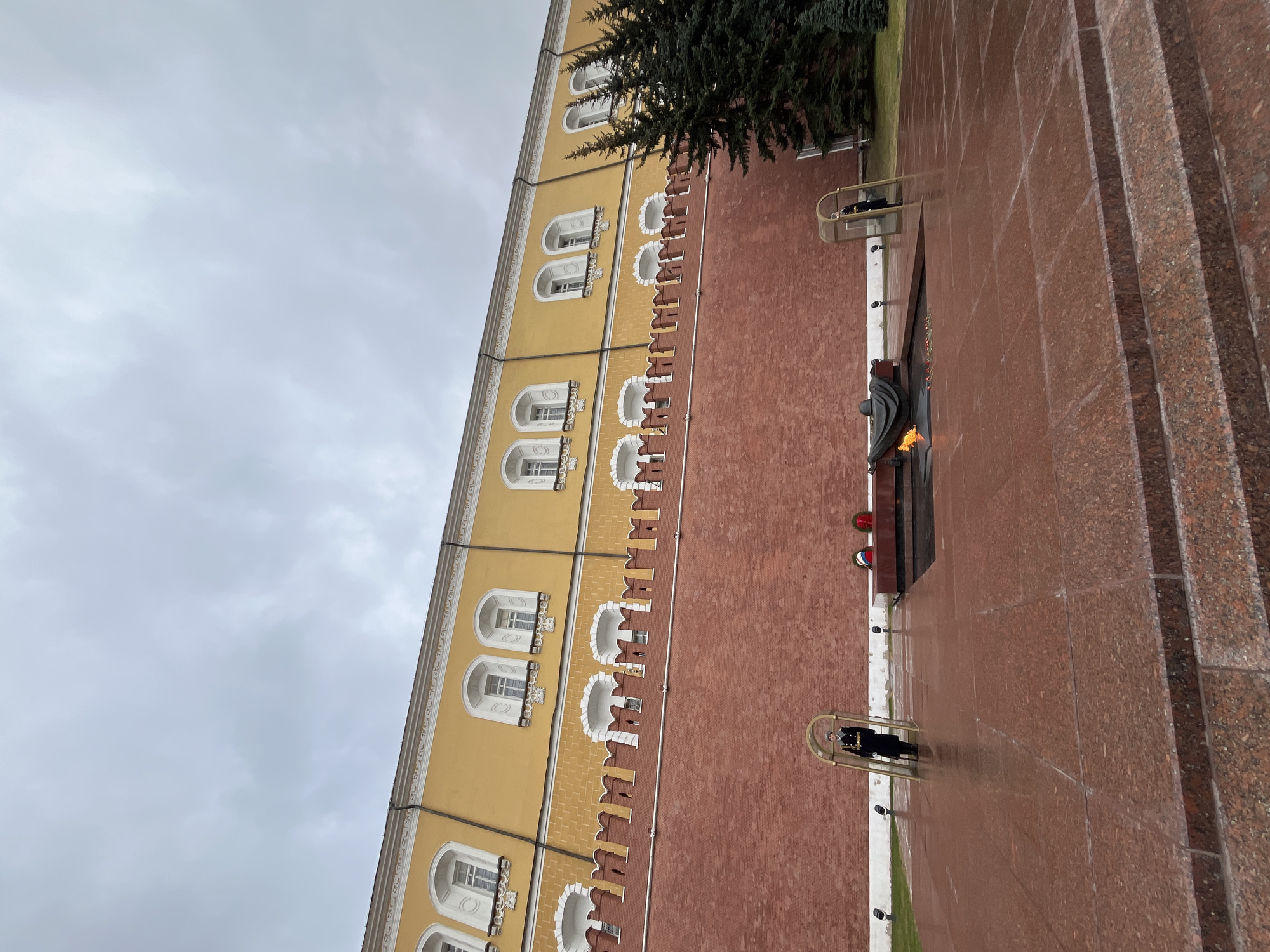 This is an eye-level, outdoor shot of the Tomb of the Unknown Soldier at the Kremlin Wall in Moscow, Russia. The wall of the building is on the left with the upper portion being yellow, and the lower portion brick red, with white architectural details around the windows. The Tomb of the Unknown Soldier is to the right with an eternal flame burning. There are two ceremonial guards on duty, standing in cages made of metal poles. The paving stones on the ground are red marble. The sky is overcast and gray.
