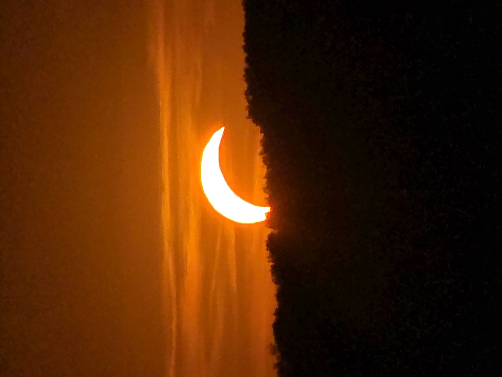 A crescent-shaped solar eclipse is visible above a dark, silhouetted mountain range under a hazy, orange sky. The sun's light filters through thin clouds, creating horizontal streaks of varying intensity. The lower portion of the image is dominated by the dark, textured silhouette of the mountain, with the bright curve of the eclipsed sun peeking over its crest.