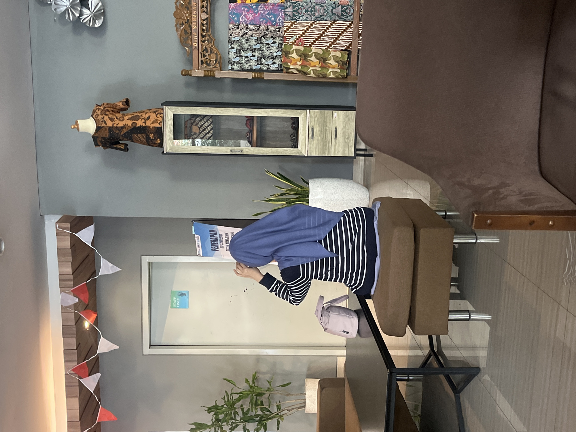 In a well-lit indoor space, likely a commercial establishment, a woman wearing a blue hijab and a black and white striped shirt is seated on a brown ottoman, her back to the camera. She appears to be adjusting her hijab with her hands. To her left, a small pink bag rests on a dark rectangular table. The environment features a grey wall with decorative elements such as a string of red and white pennant flags and a mannequin displaying a brown and black patterned garment. Various plants in white pots add greenery to the scene. A tall, narrow cabinet with glass doors and drawers stands to the right of the mannequin, and a wooden display rack showcasing colorful fabrics is positioned further to the right. In the background, a closed door is visible with a "STAFF ONLY" sign. The flooring is tiled. This scene is situated in the city of Gapurosukolilo, Indonesia.