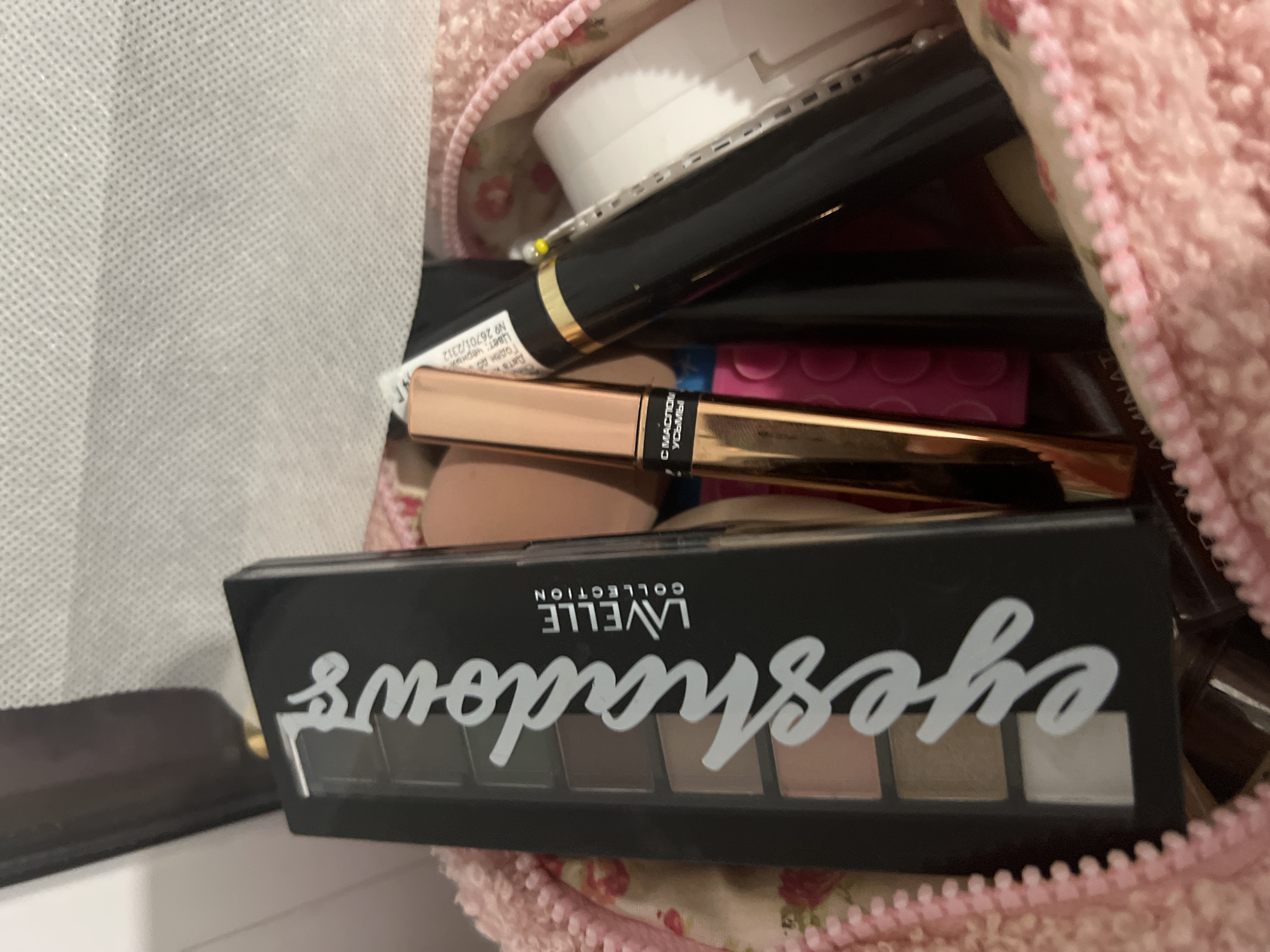 The image shows a close-up of a pink makeup bag, partially unzipped, revealing an assortment of cosmetics. Dominating the foreground is a black eyeshadow palette with the word "eyeshadow" written vertically in white script. Several shades of eyeshadow are visible within the palette. To the right of the eyeshadow palette, a rose-gold colored tube, possibly lipstick or gloss, is positioned. Further to the right, a black mascara tube with a gold band is visible, along with other makeup items. The background consists of the textured interior of the makeup bag and a white surface that appears to be part of a larger object, perhaps a travel case or a vanity. There are no people or any recognizable landmarks of Yekaterinburg, Russia in the image.